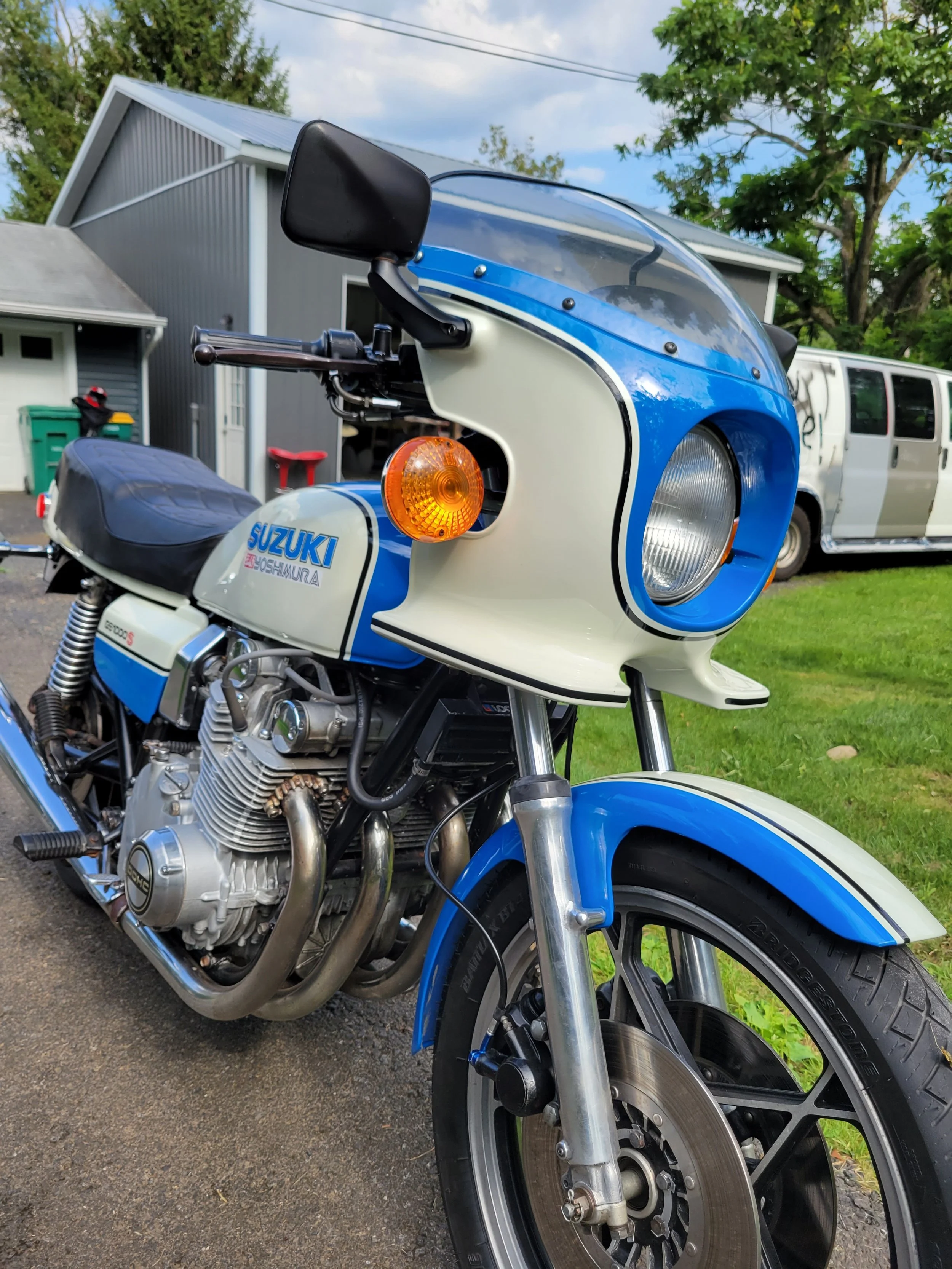 A vintage blue and white Suzuki motorcycle parked on a driveway in front of houses with green lawns, trees, and a vintage van in the background.