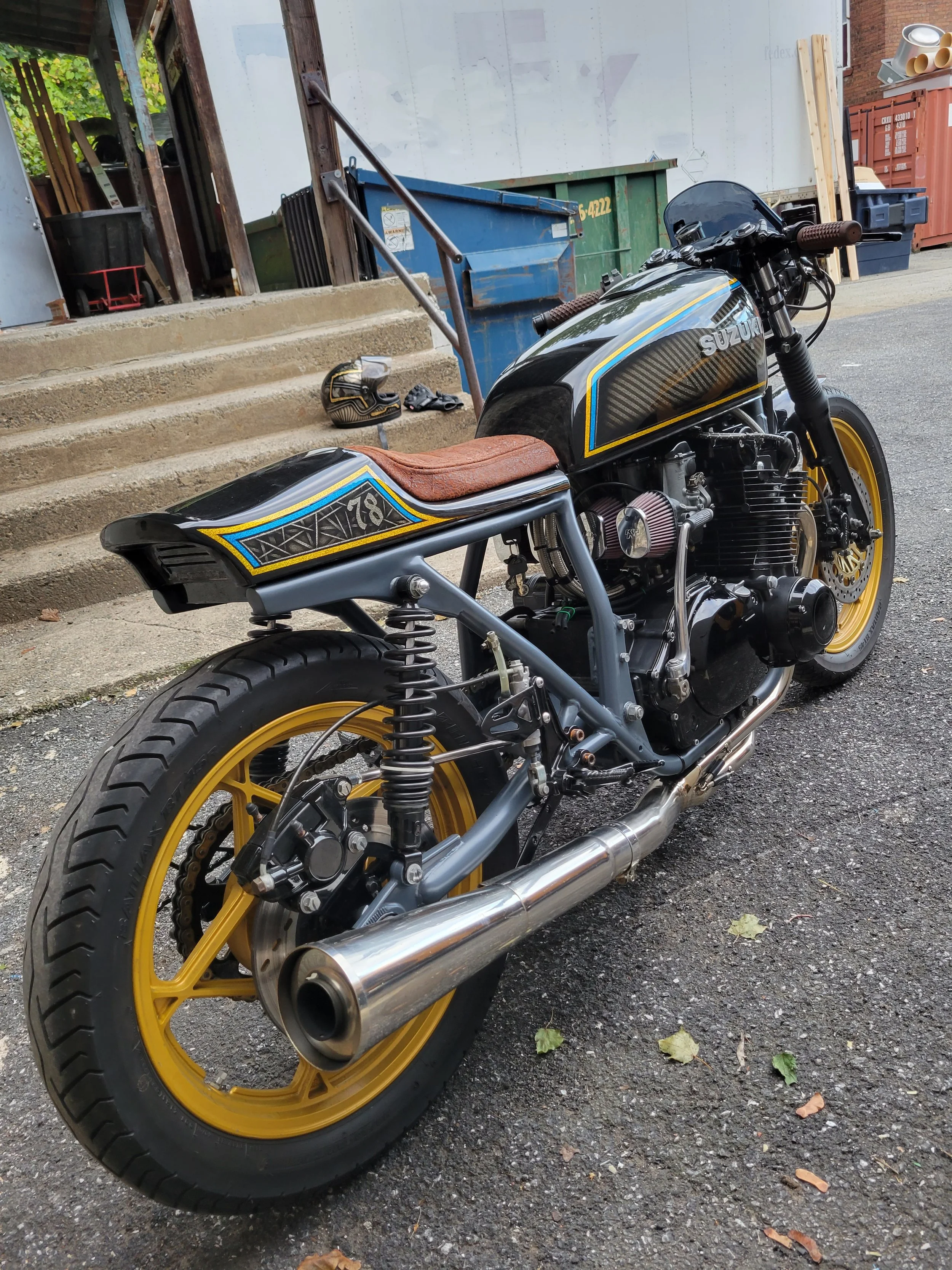 A custom black and gold Suzuki motorcycle with a brown leather seat, parked on a gravel surface in front of building stairs, with a helmet on the ground nearby.