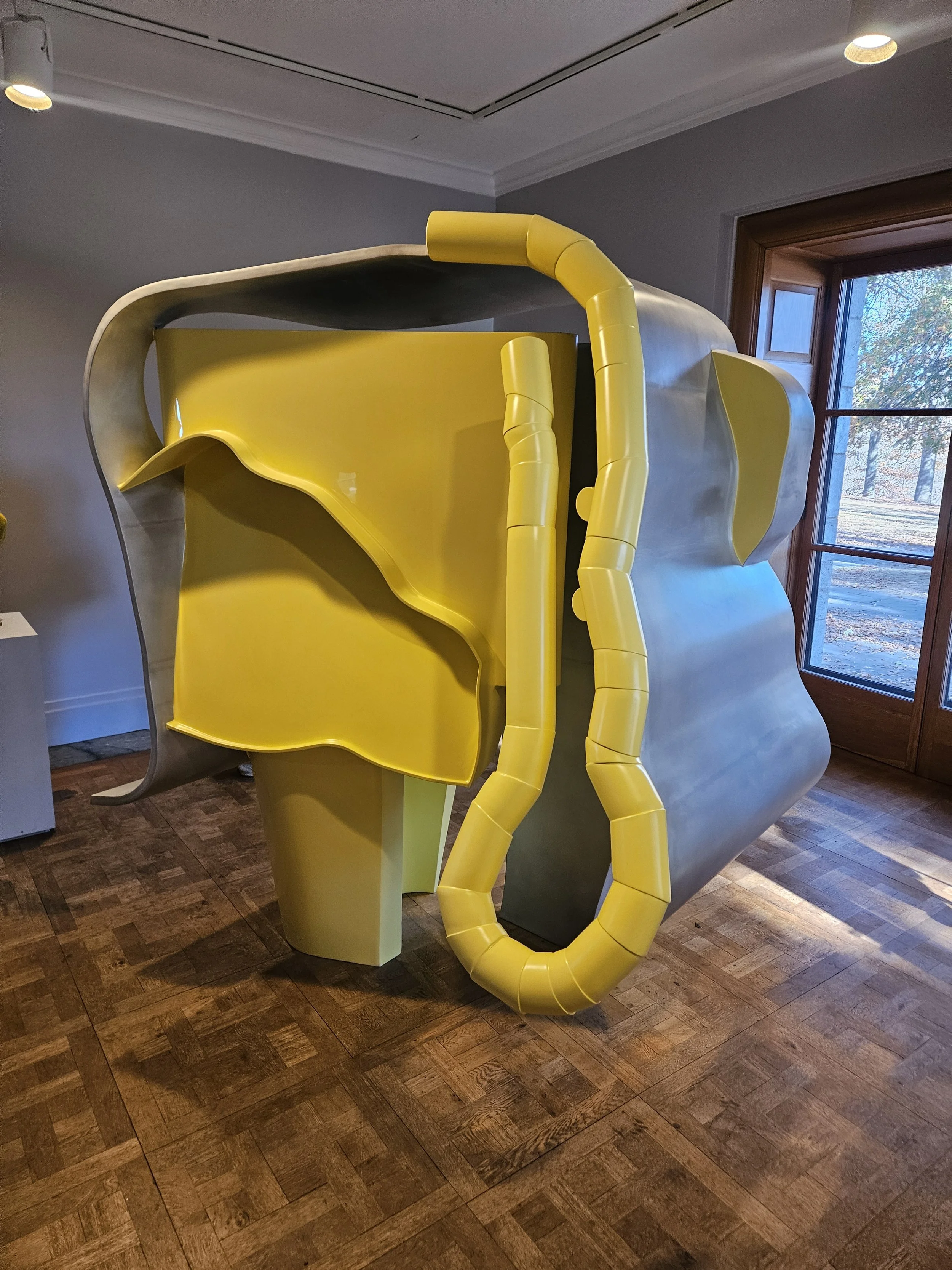 Yellow and gray abstract sculpture with curvy and tubular shapes inside a room near a window.