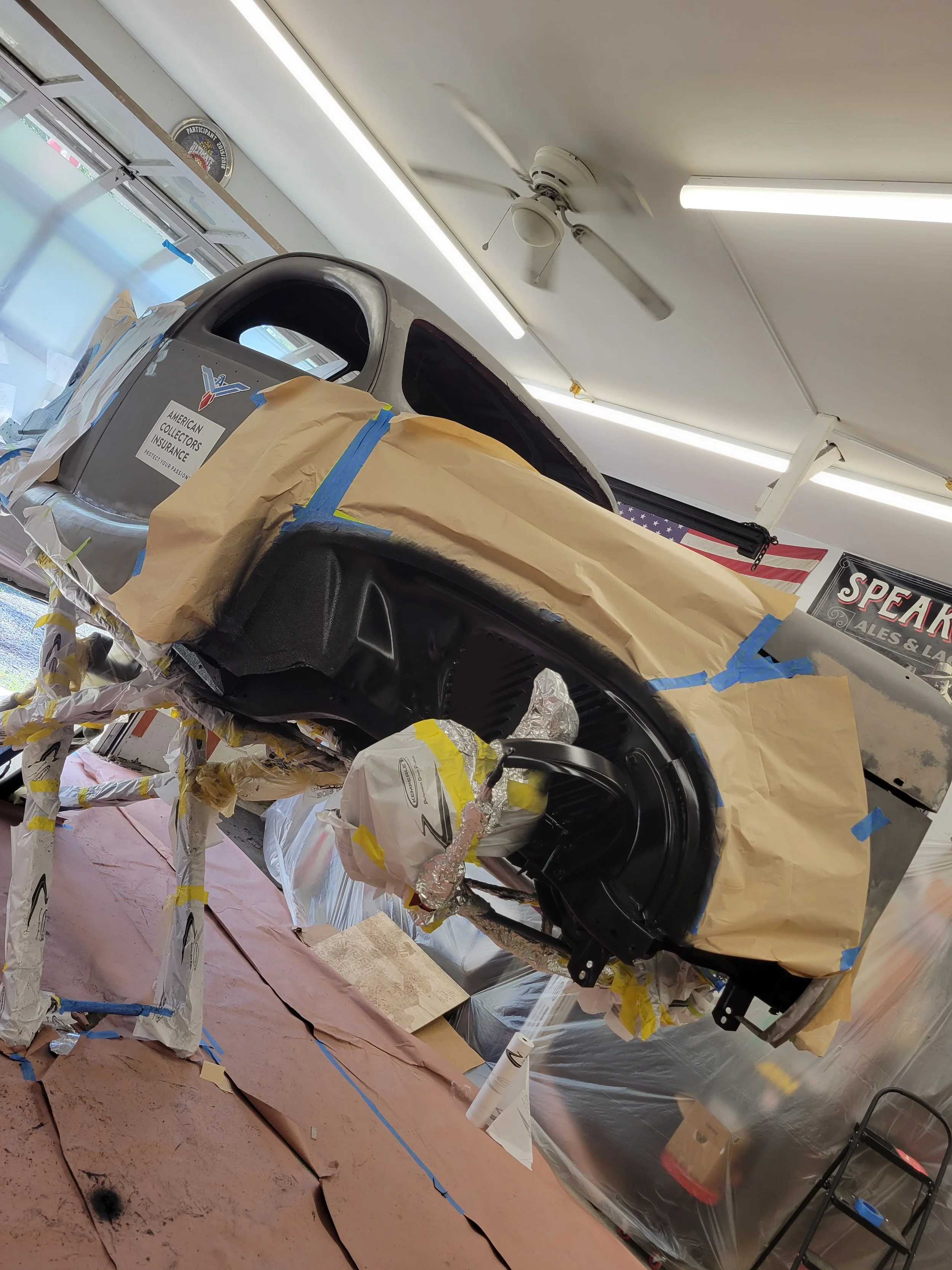 A car undergoing restoration, suspended on a makeshift frame in a workshop. The car's body is partially masked with paper and tape, with some components covered in foil or plastic for painting or repair.