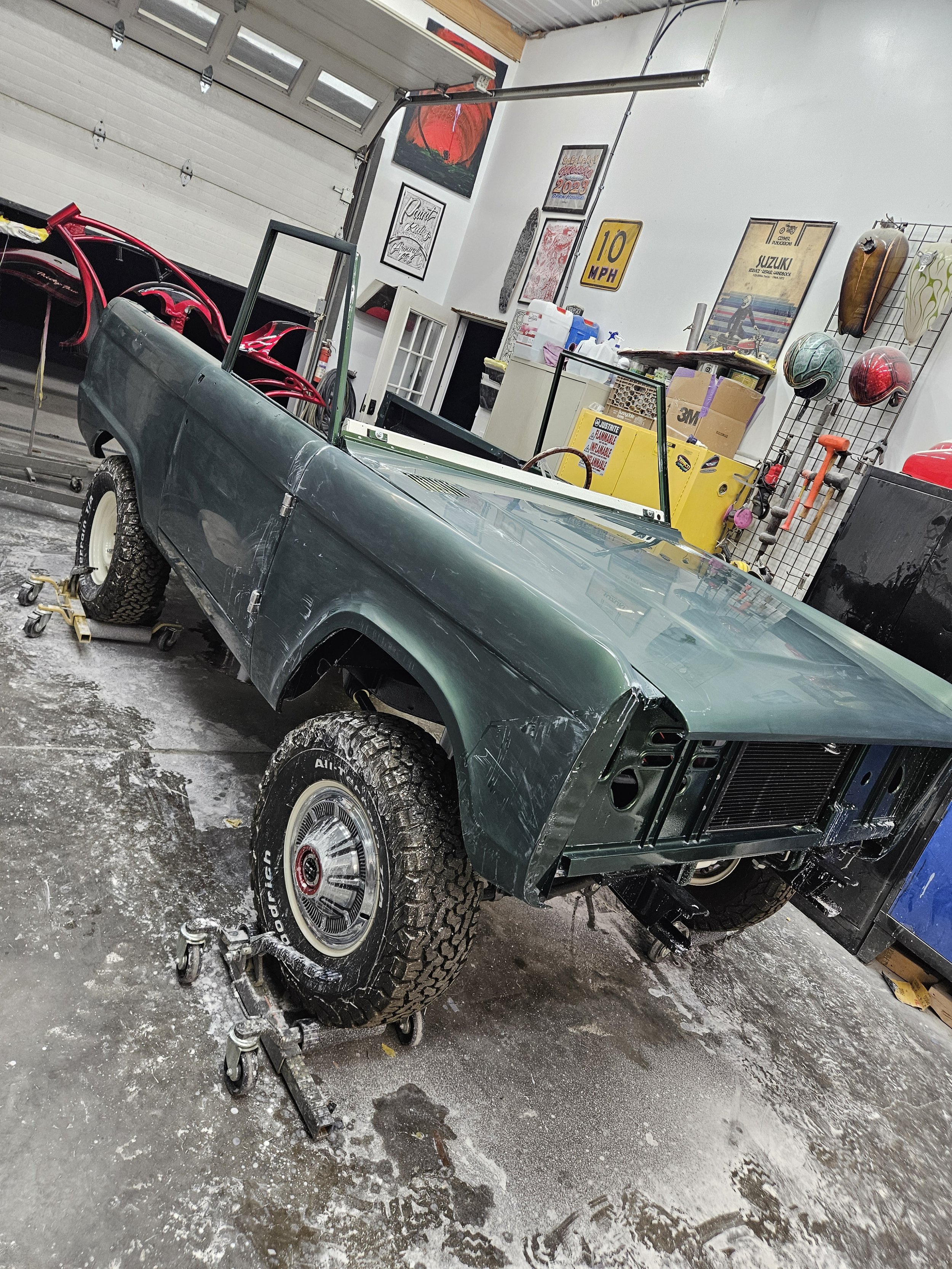 A vintage green pickup truck in a garage undergoing restoration, with wheels and parts removed, surrounded by tools, helmets, and garage decor.