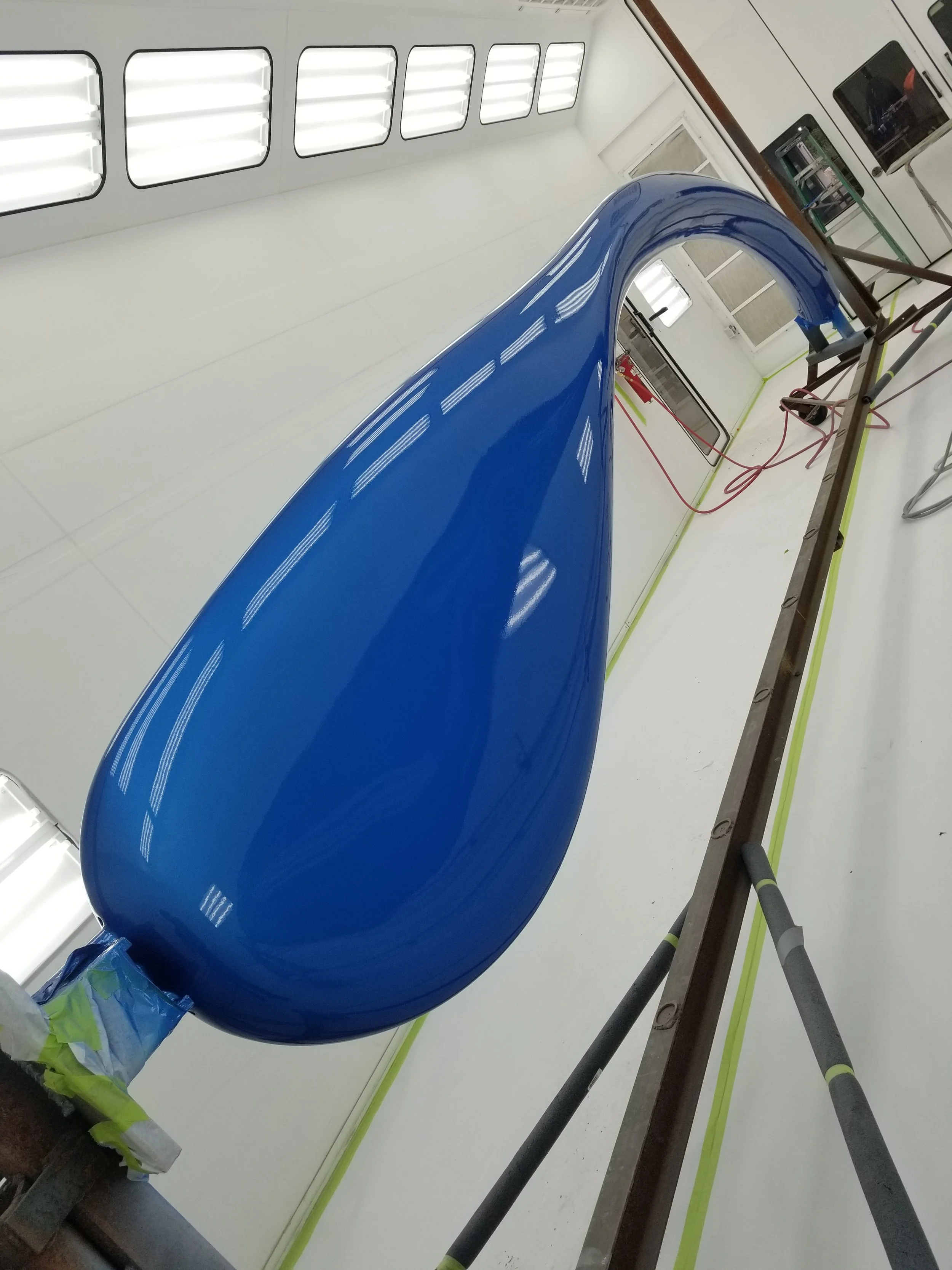 A large, shiny, blue teardrop-shaped object in a bright white room, possibly an aircraft nose cone or a similar component, hanging and being painted or coated.