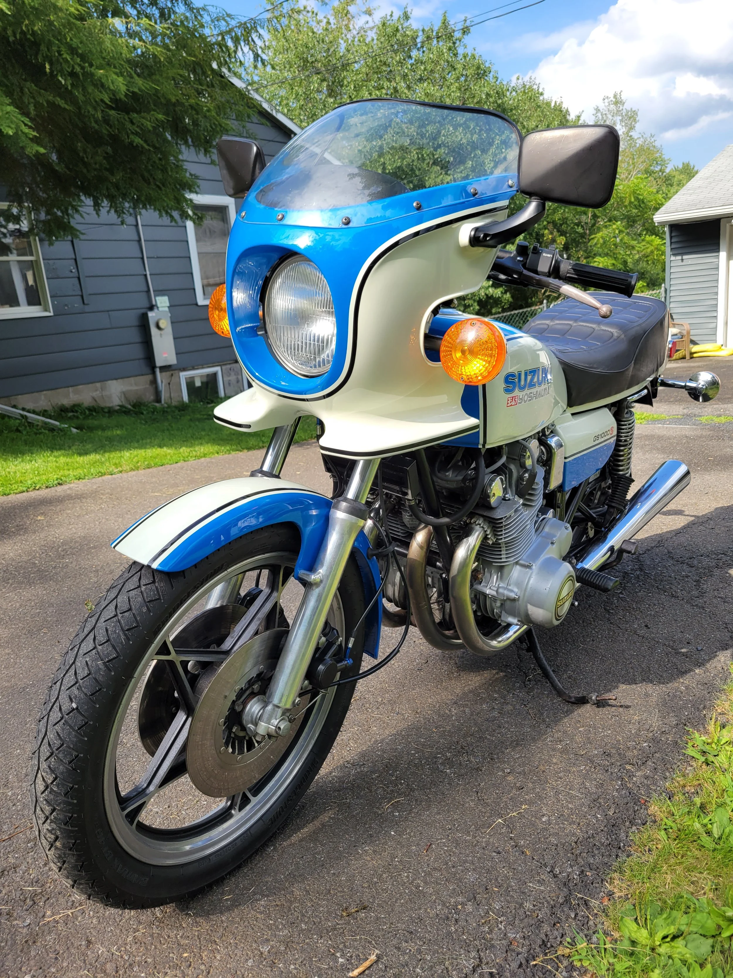 A vintage Suzuki motorcycle with a blue and white color scheme parked on a driveway in front of a house with blue siding and green trees.
