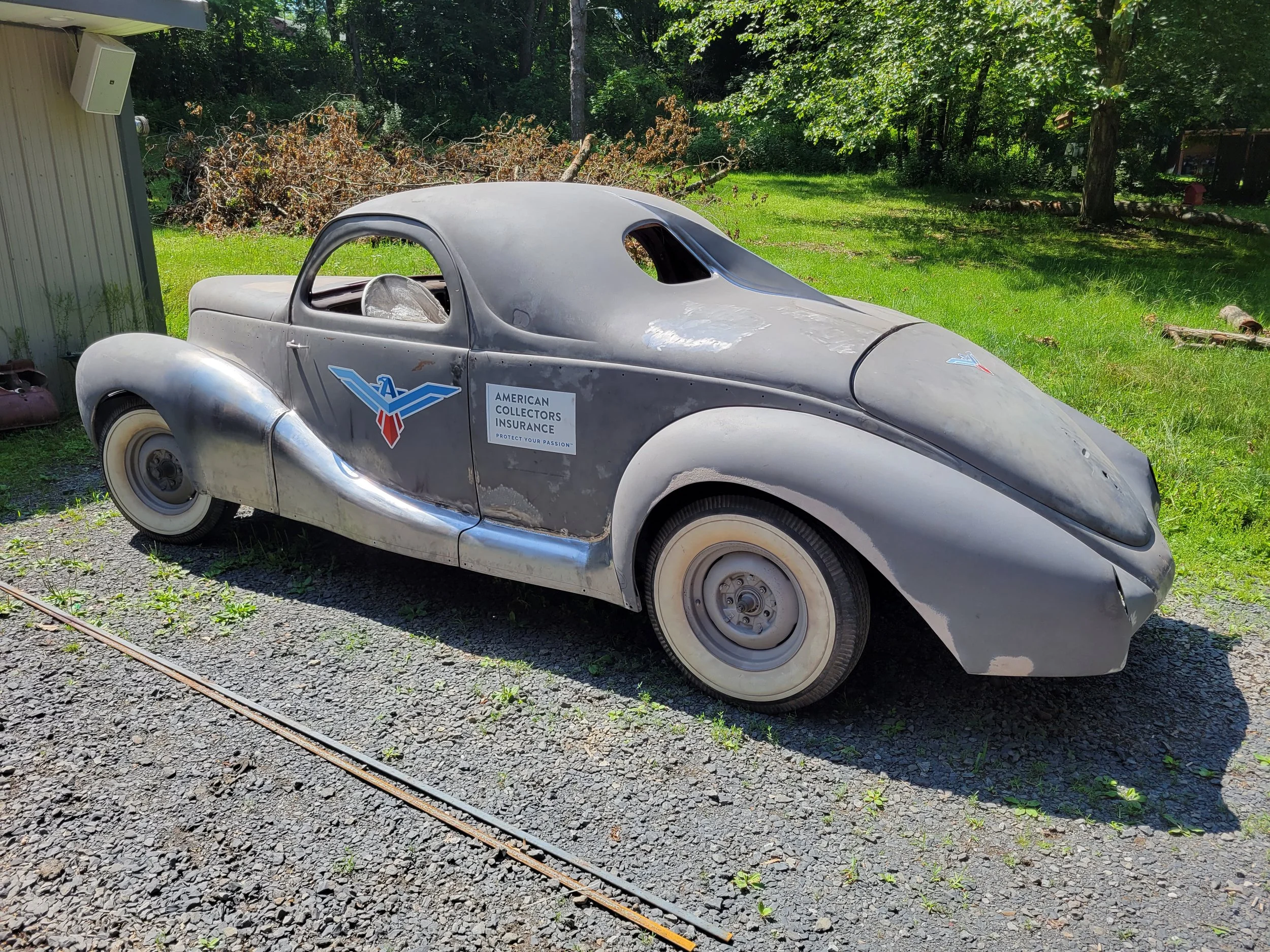 An old, weathered vintage car with rounded body, worn paint, and a logo of American Collectors Insurance on its door, parked on a gravel surface. The car is dusty and appears to be in storage or abandoned, with a grassy background and trees.
