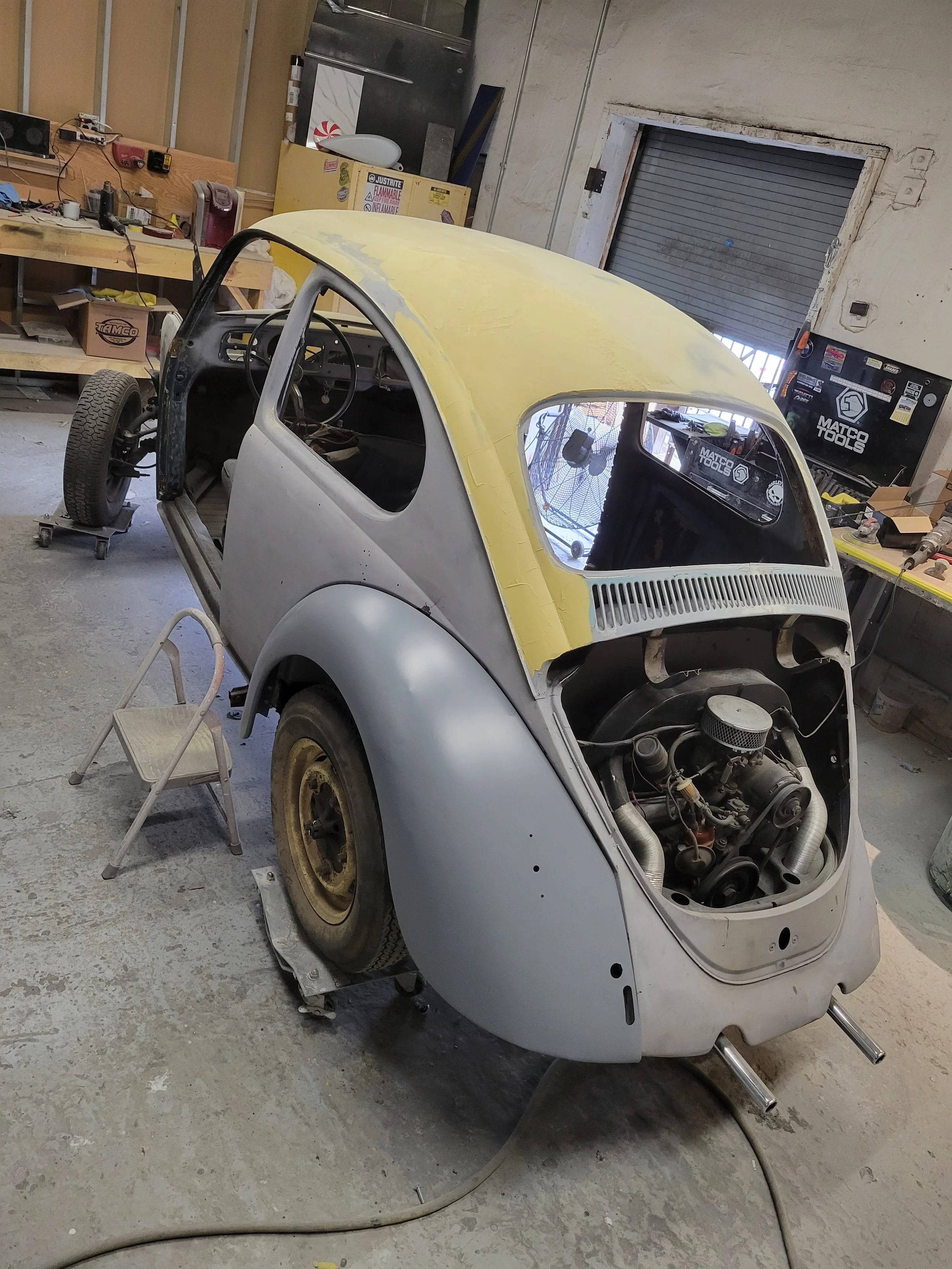 Restoration of a small vintage car in a workshop, partially assembled with the body fitted over the engine and wheels, with workbenches and tools in the background.