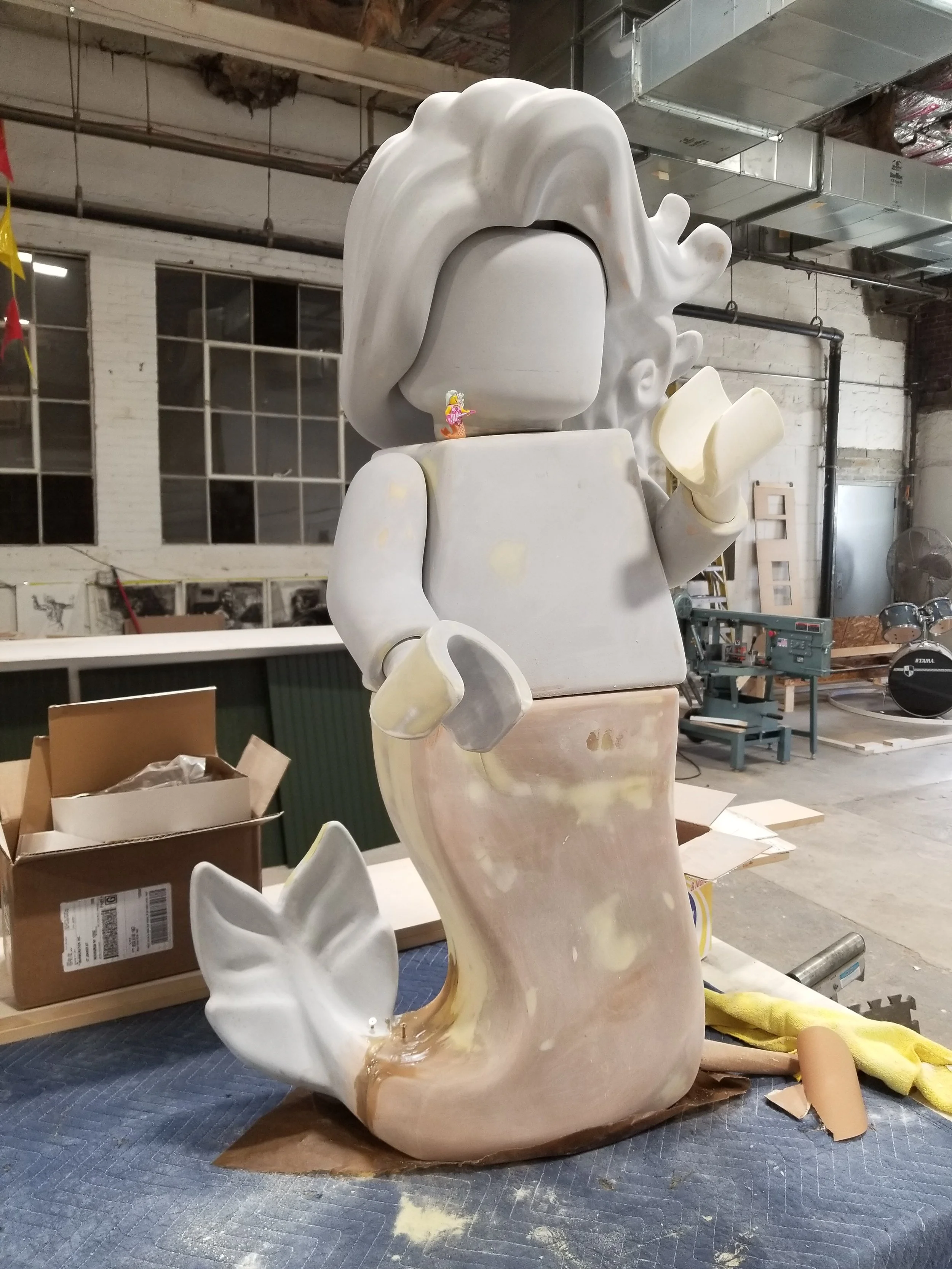 A large, unfinished sculpture of a LEGO mermaid sculpture with big hair in a workshop with tools and materials around.