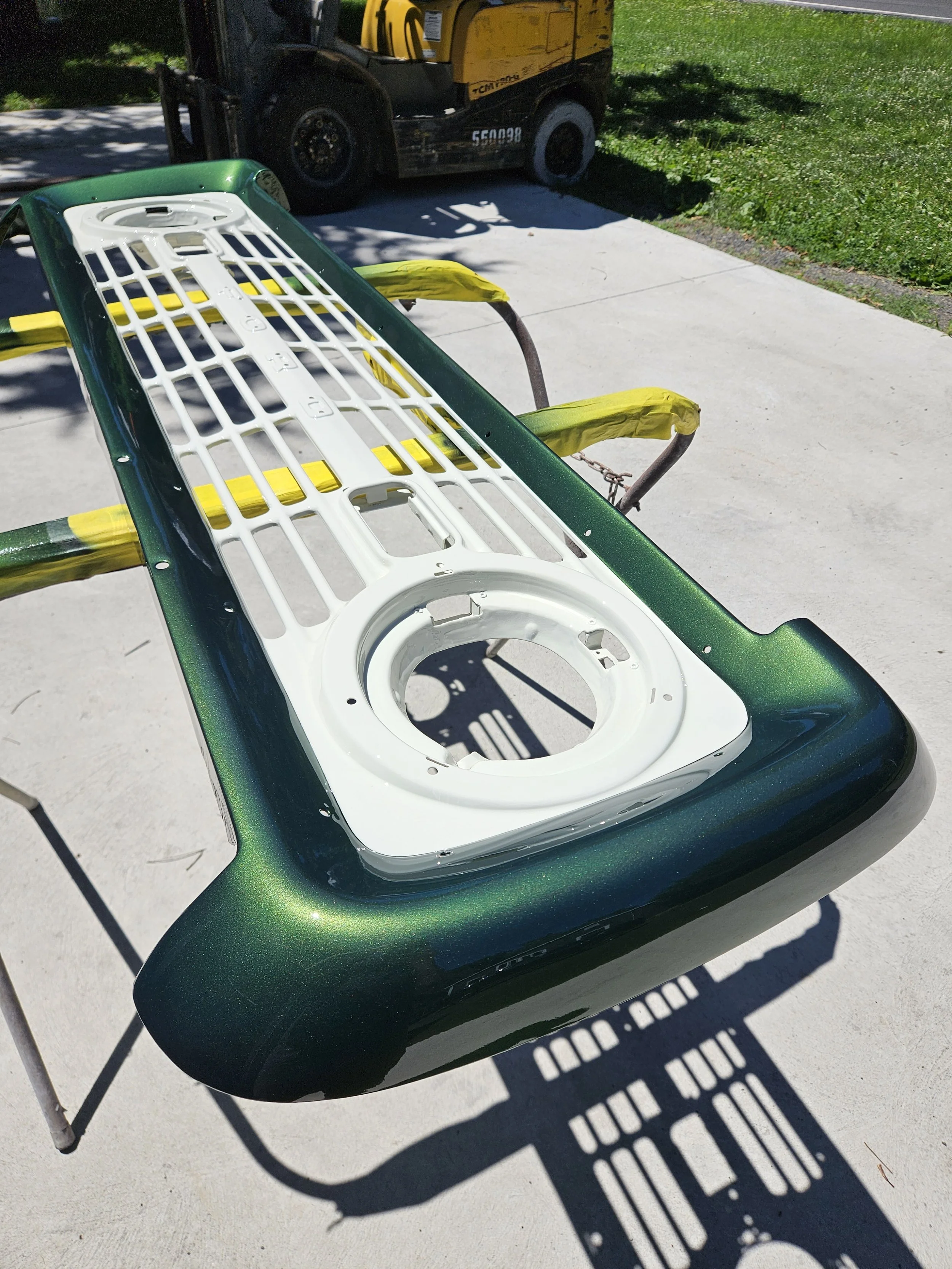 A painted skateboard deck with a green and black sparkly finish, placed on a work stand outdoors. The skateboard's top side is visible, showing a white grip tape and mounting hardware in the center. In the background, there is a yellow and black piec