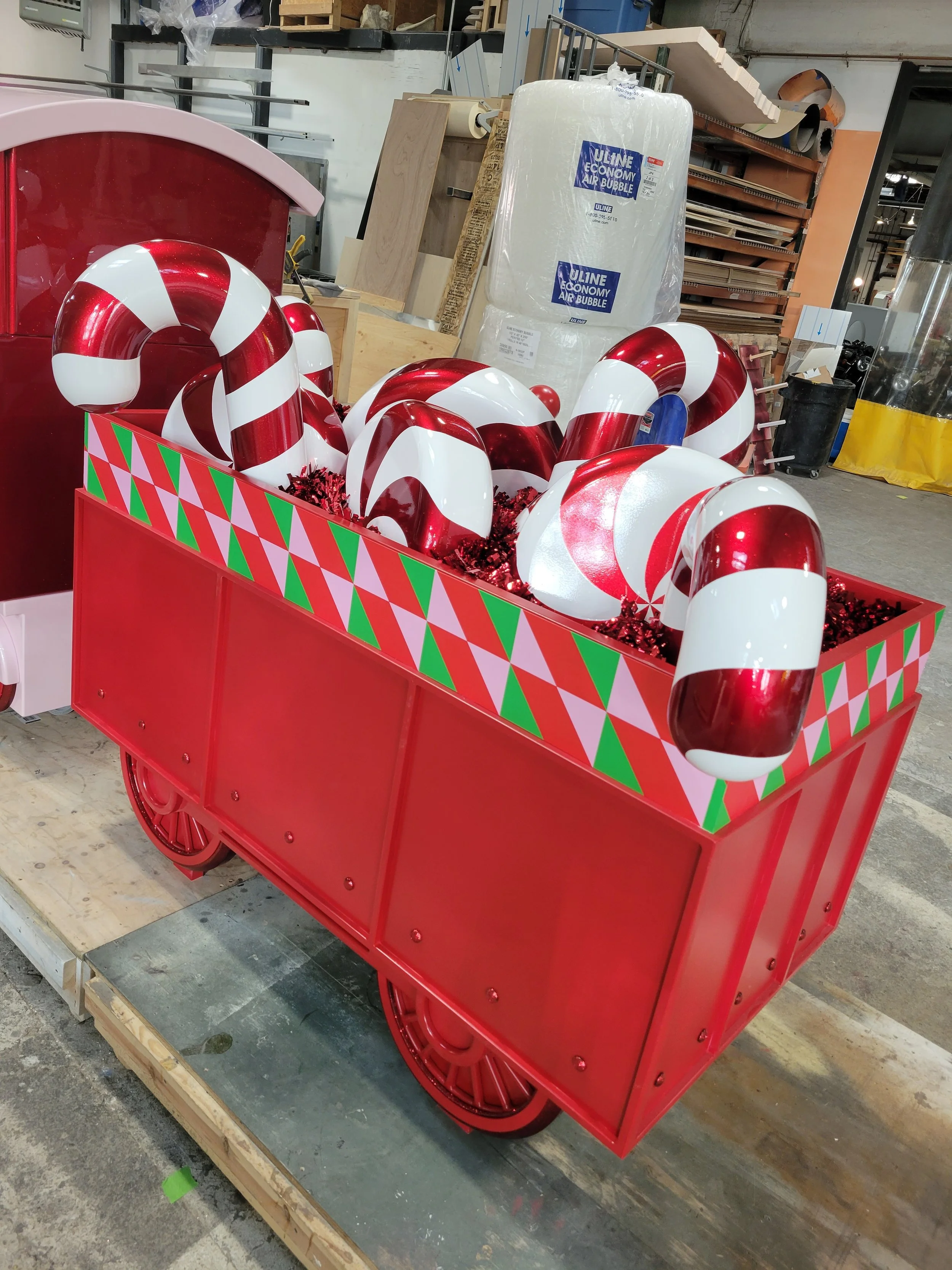 A red shopping cart decorated with a green, red, and pink harlequin pattern on its sides, filled with large candy cane ornaments with red and white stripes inside a warehouse or workshop setting.