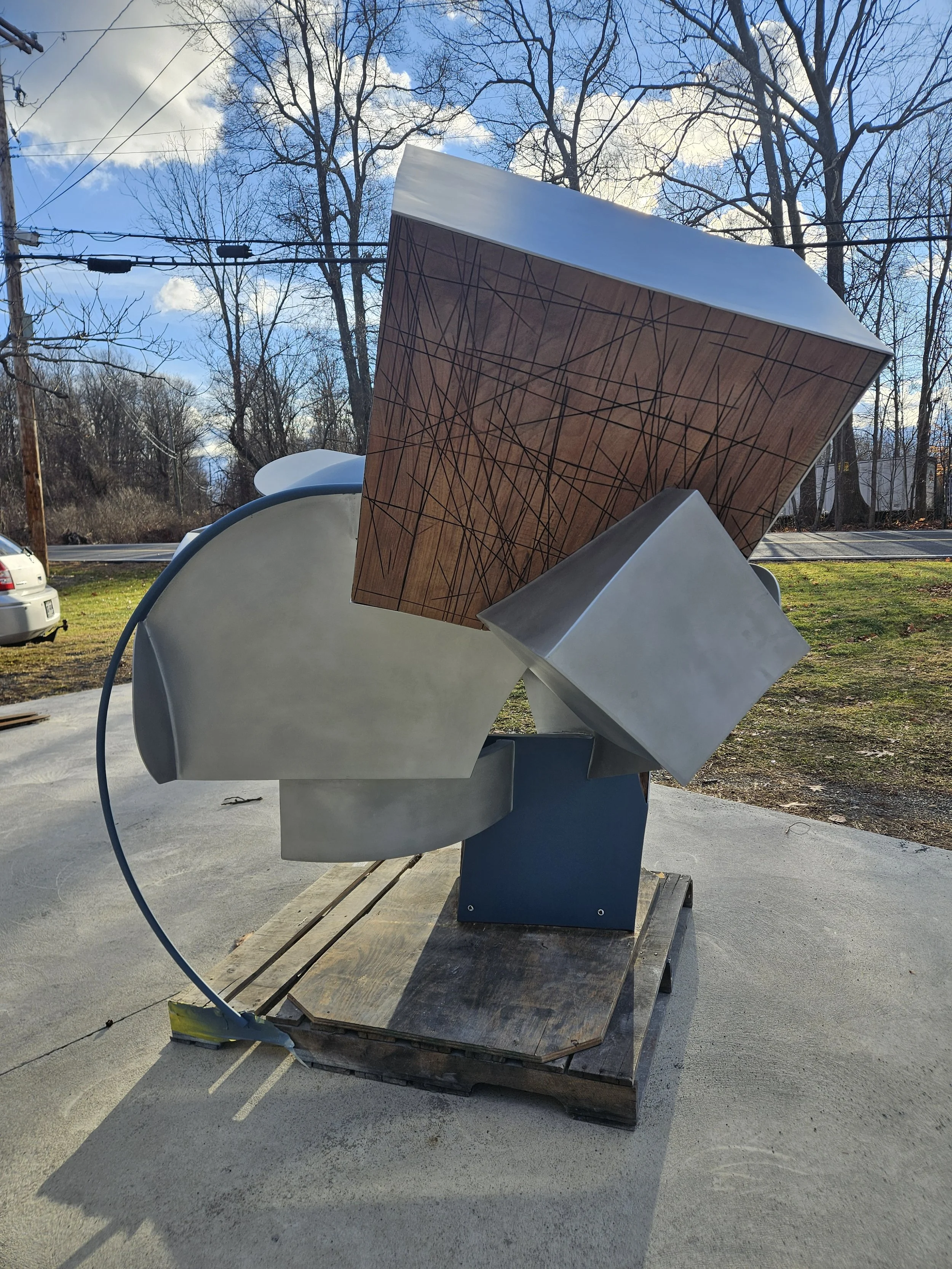 Abstract metal sculpture with geometric forms and wooden panel, positioned outdoors on a wooden pallet, with trees, power lines, and a car in the background.
