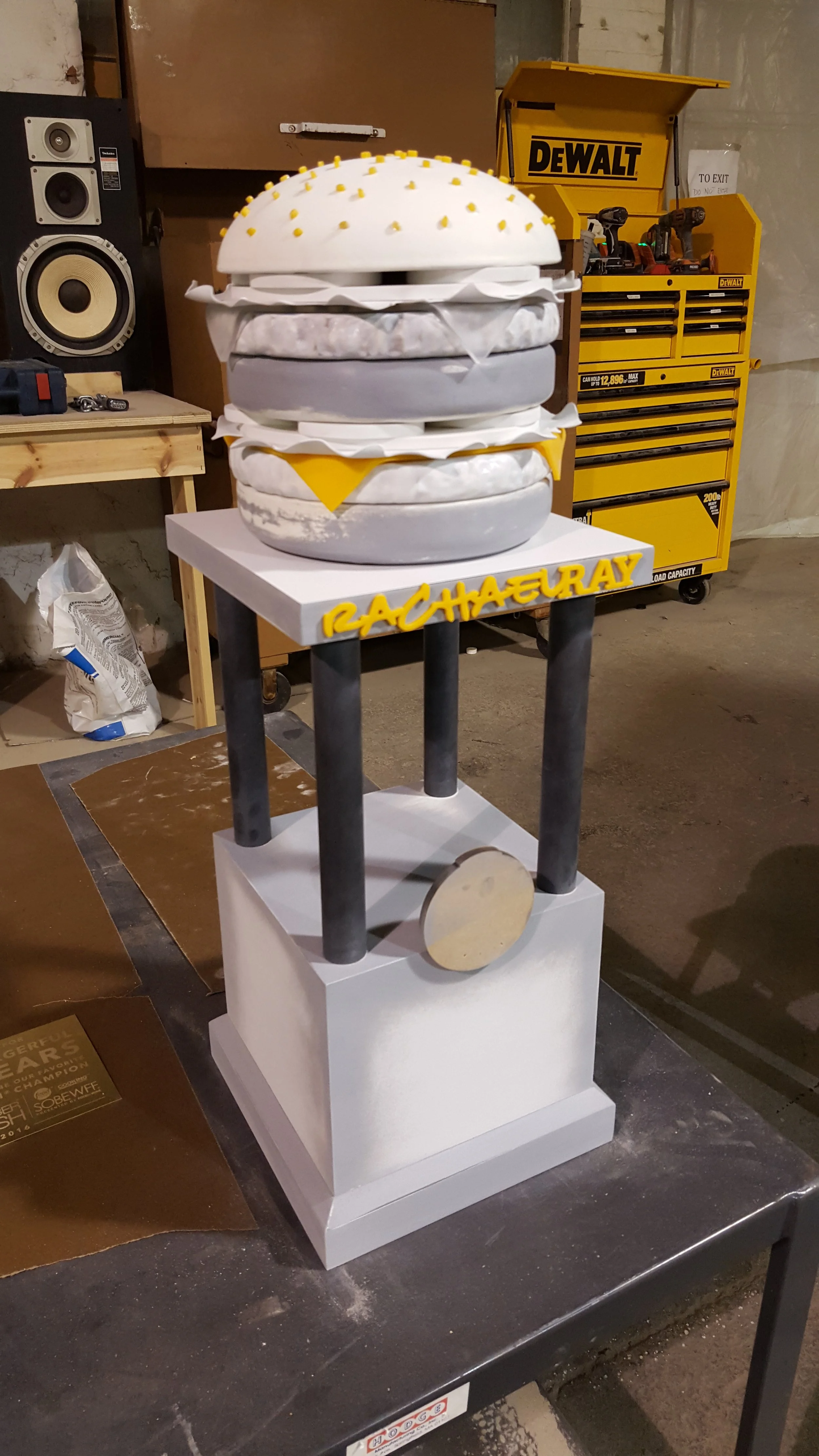 A multi-layered burger-shaped sculpture trophy inside a workshop, with a yellow Dewalt tool chest in the background, and the name 'Rachael Ray' displayed