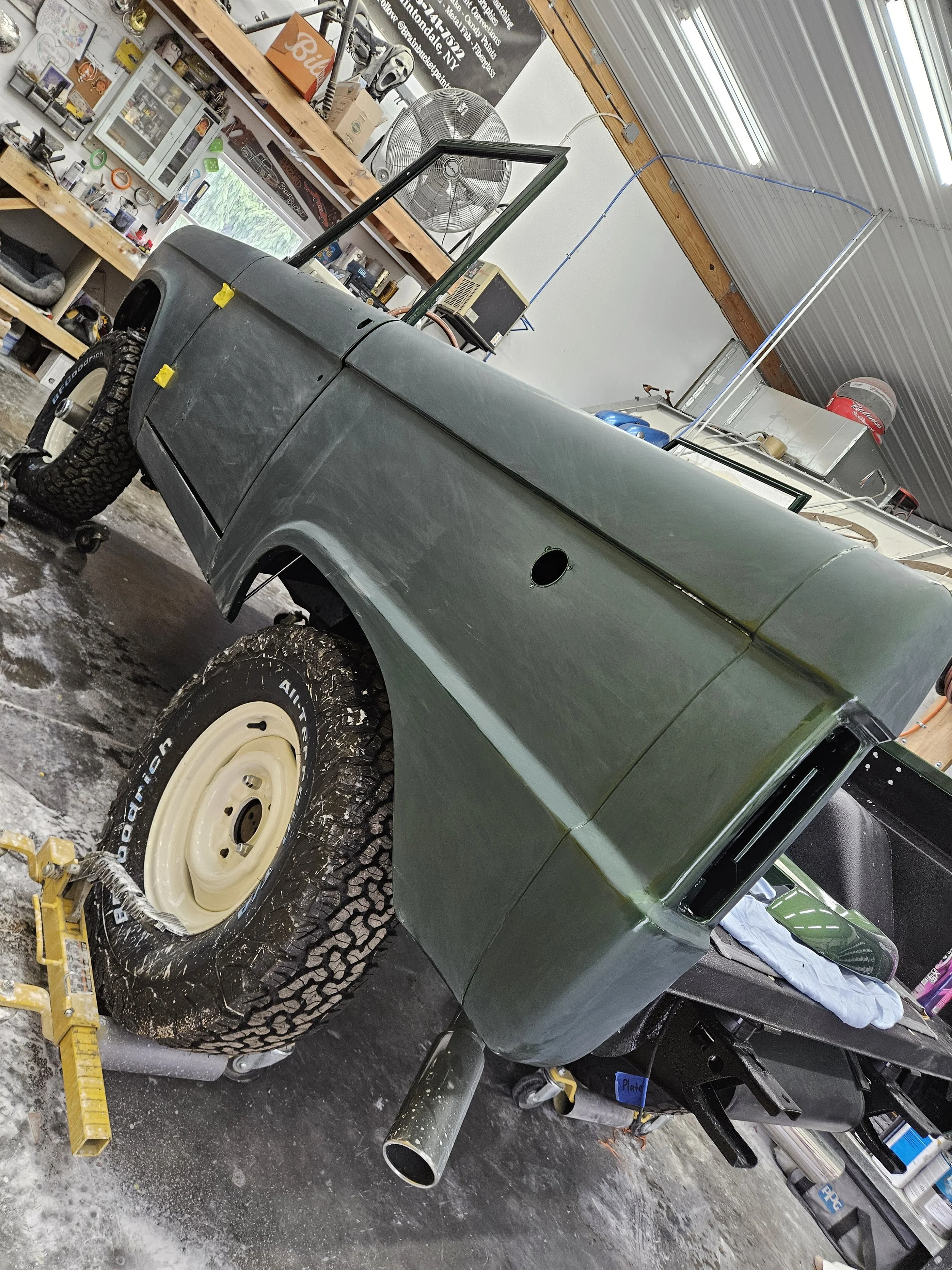 An unfinished vintage amphibious vehicle in a garage, with a custom-built frame, large off-road tires, and a glass windshield. The garage has shelves with various tools and supplies.