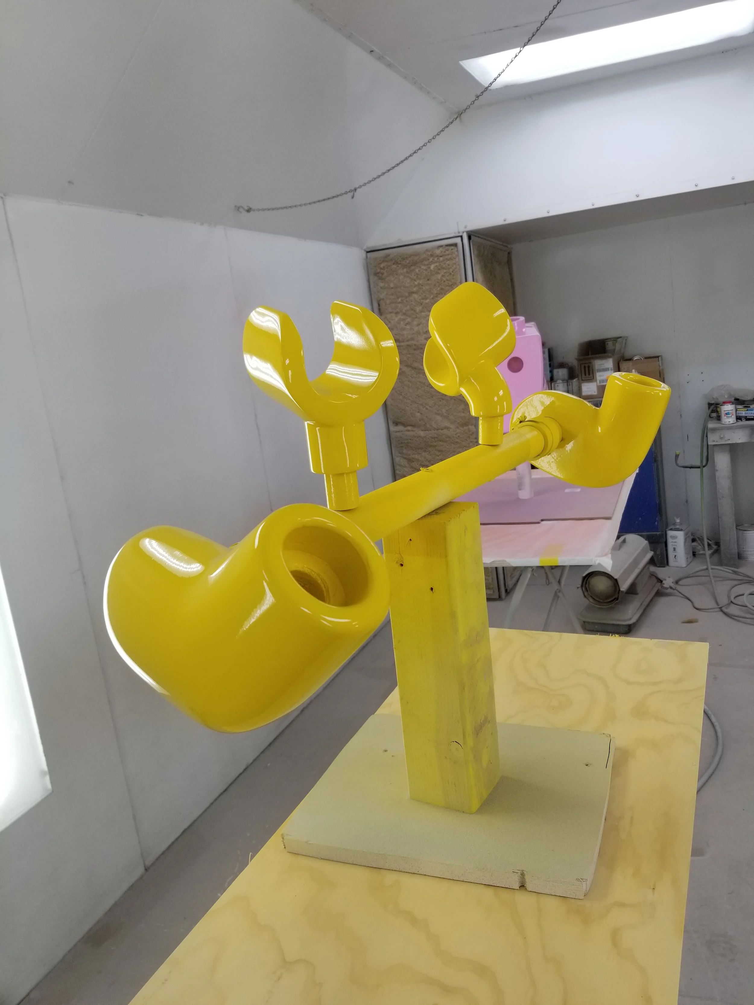 Bright yellow sculptural artwork resembling a robotic face with a paddle, a music note, and a pipe, mounted on a yellow wooden base in an indoor workshop.
