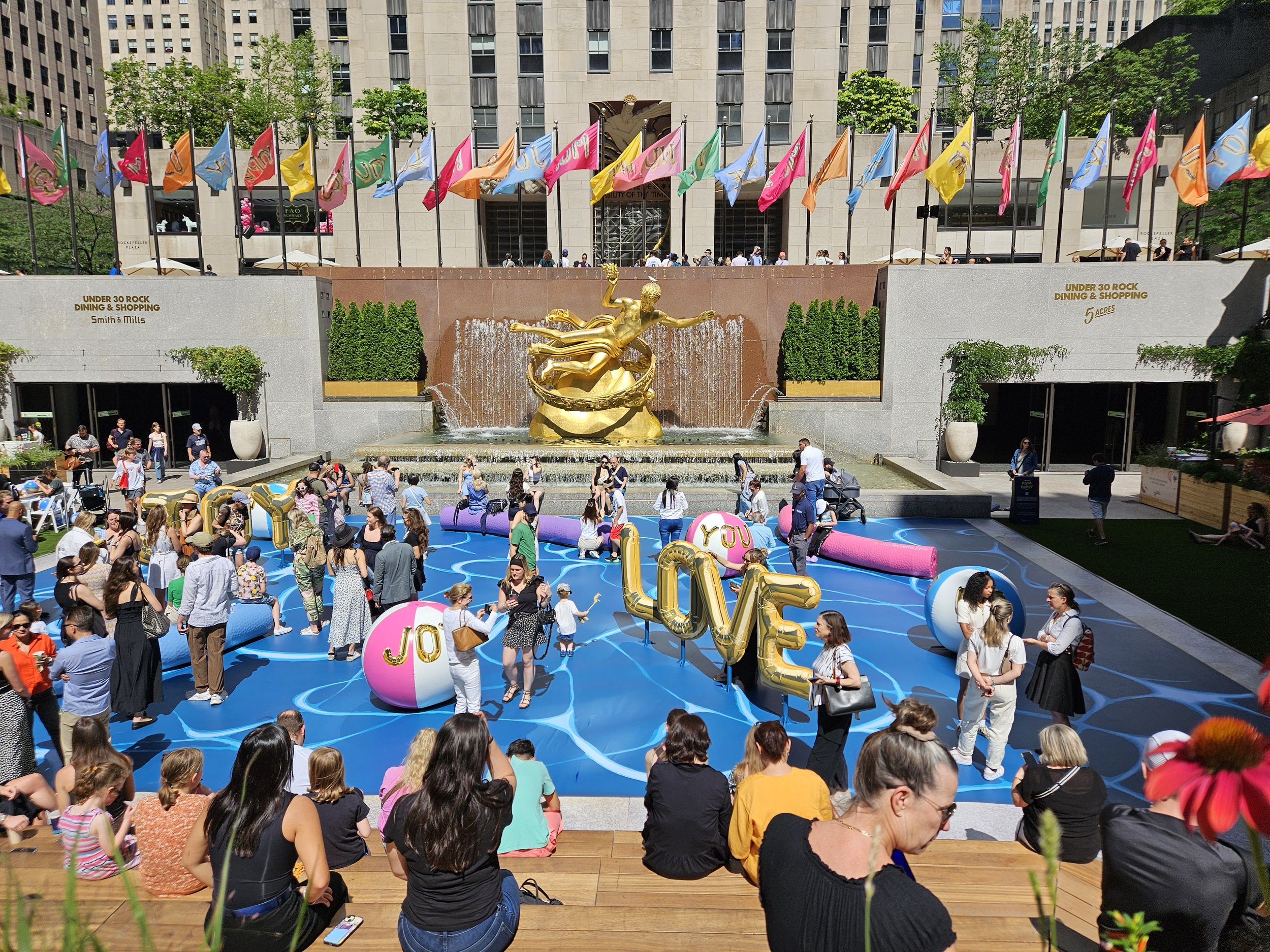 Crowd gathered in front of a fountain with gold sculptures and colorful flags, some people sitting on wooden benches and others walking around, celebrating at an outdoor event in a city square.