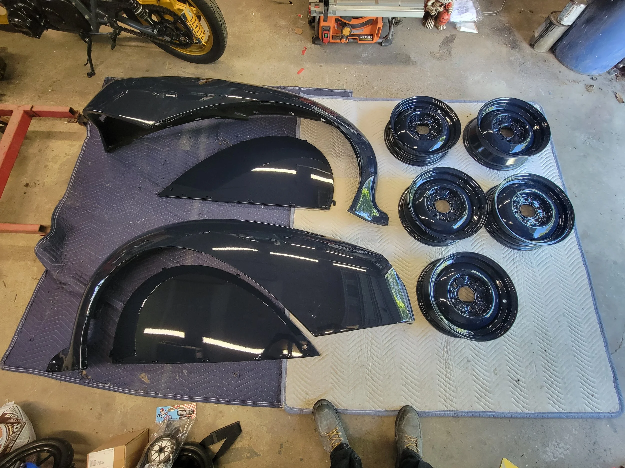 Black car fender and wheel rims placed on a quilted surface in a garage. The fender appears to be newly painted, and the rims are stacked in a group of six.