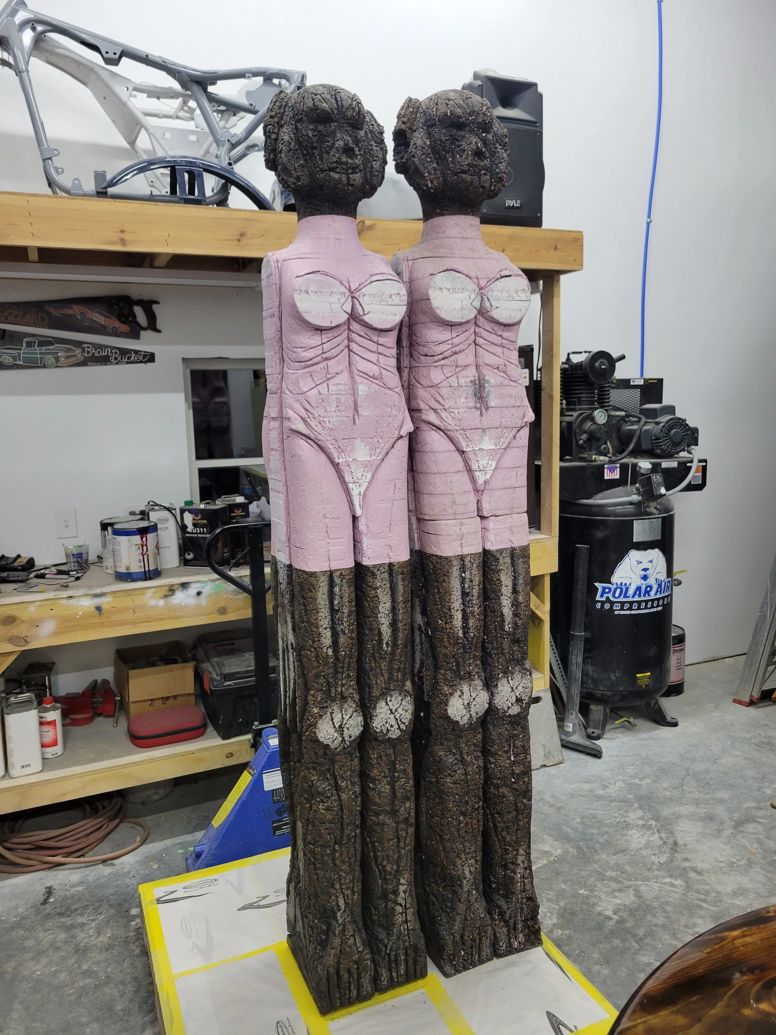 Two wood sculptures of figures with dark, textured heads and pink painted torsos, standing side by side in a workshop.