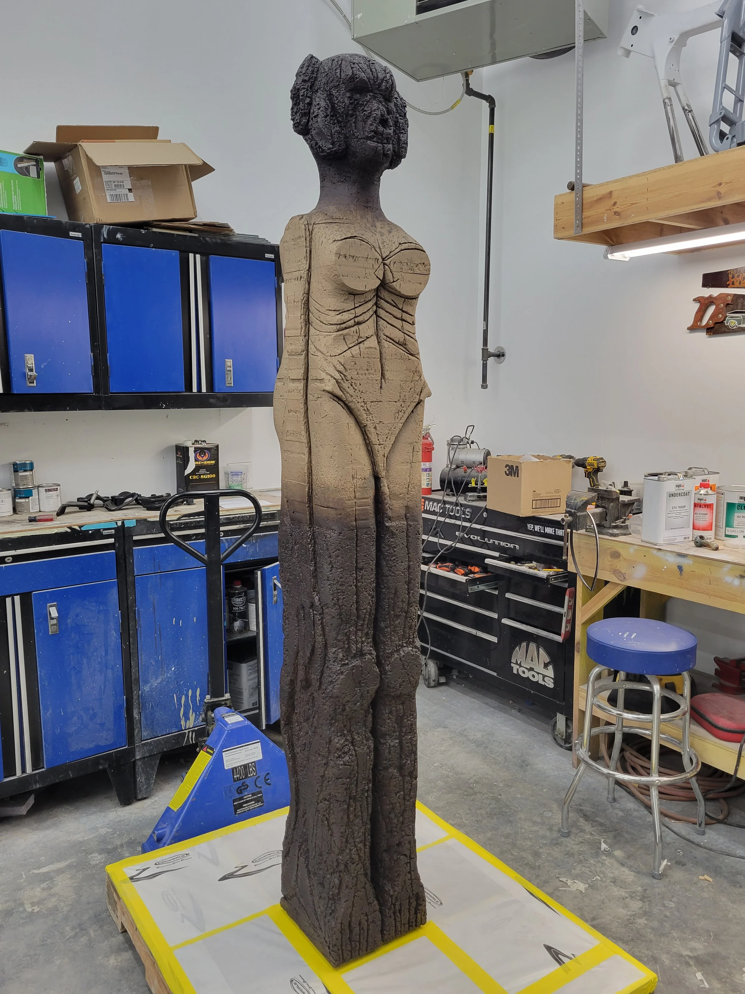 Wooden sculpture of a woman with a detailed hairstyle, standing upright in a workshop with tools and storage cabinets around.