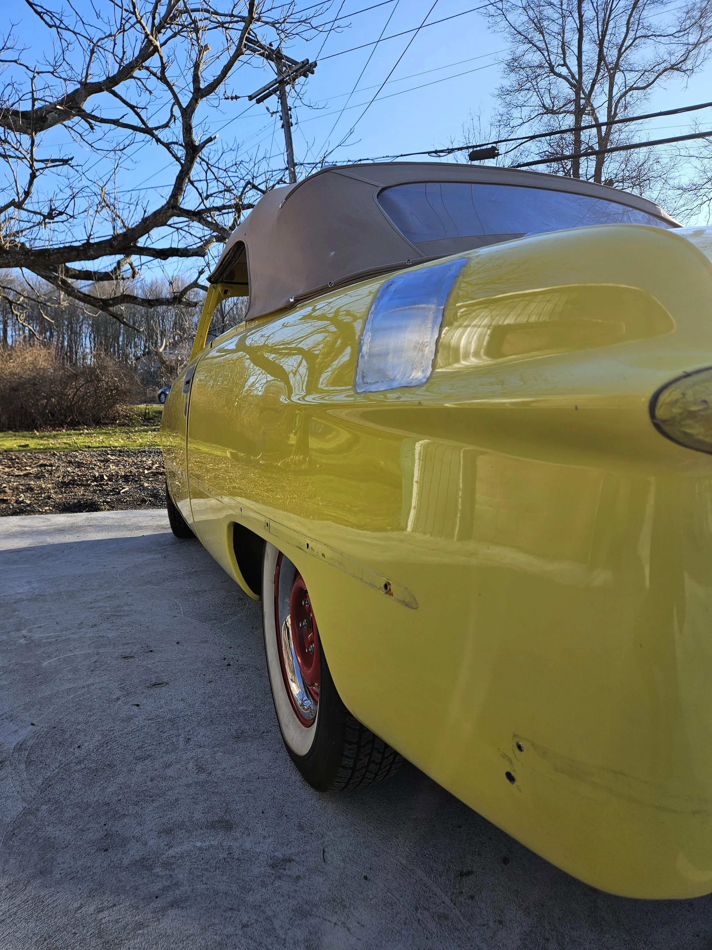 A yellow convertible car with a brown soft top, parked on a concrete surface outdoors. The background shows leafless trees, power lines, and a clear blue sky.