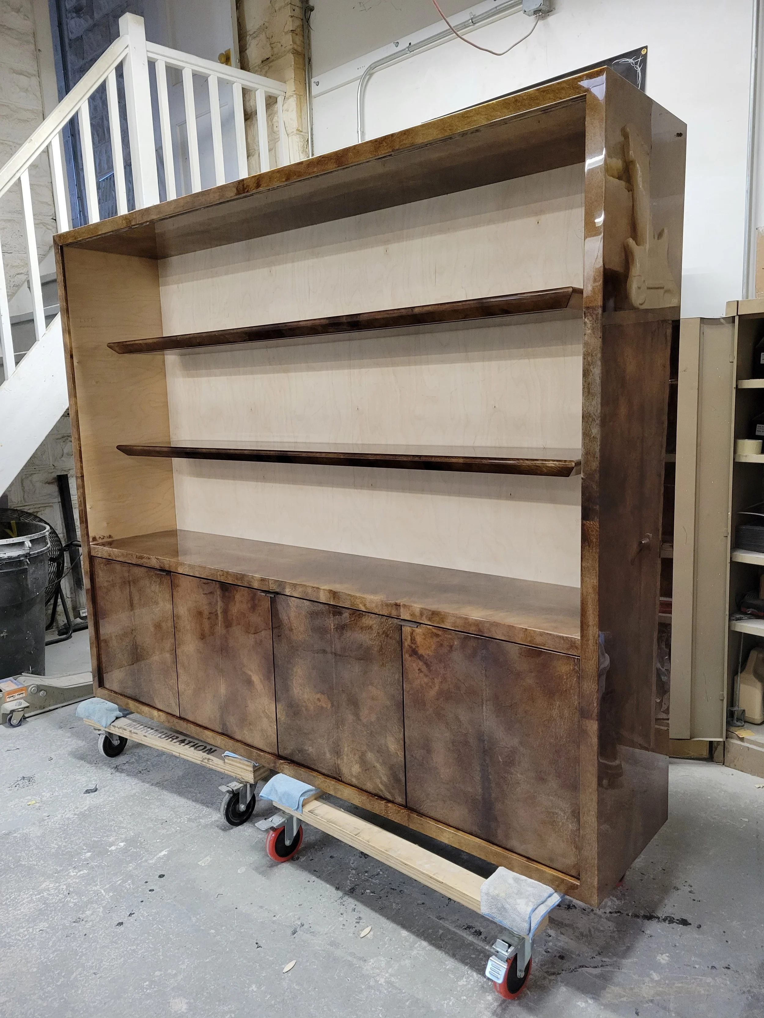 A large wooden display cabinet with three open shelves and four closed doors at the bottom. It is on a wheeled platform in a workshop or warehouse setting.
