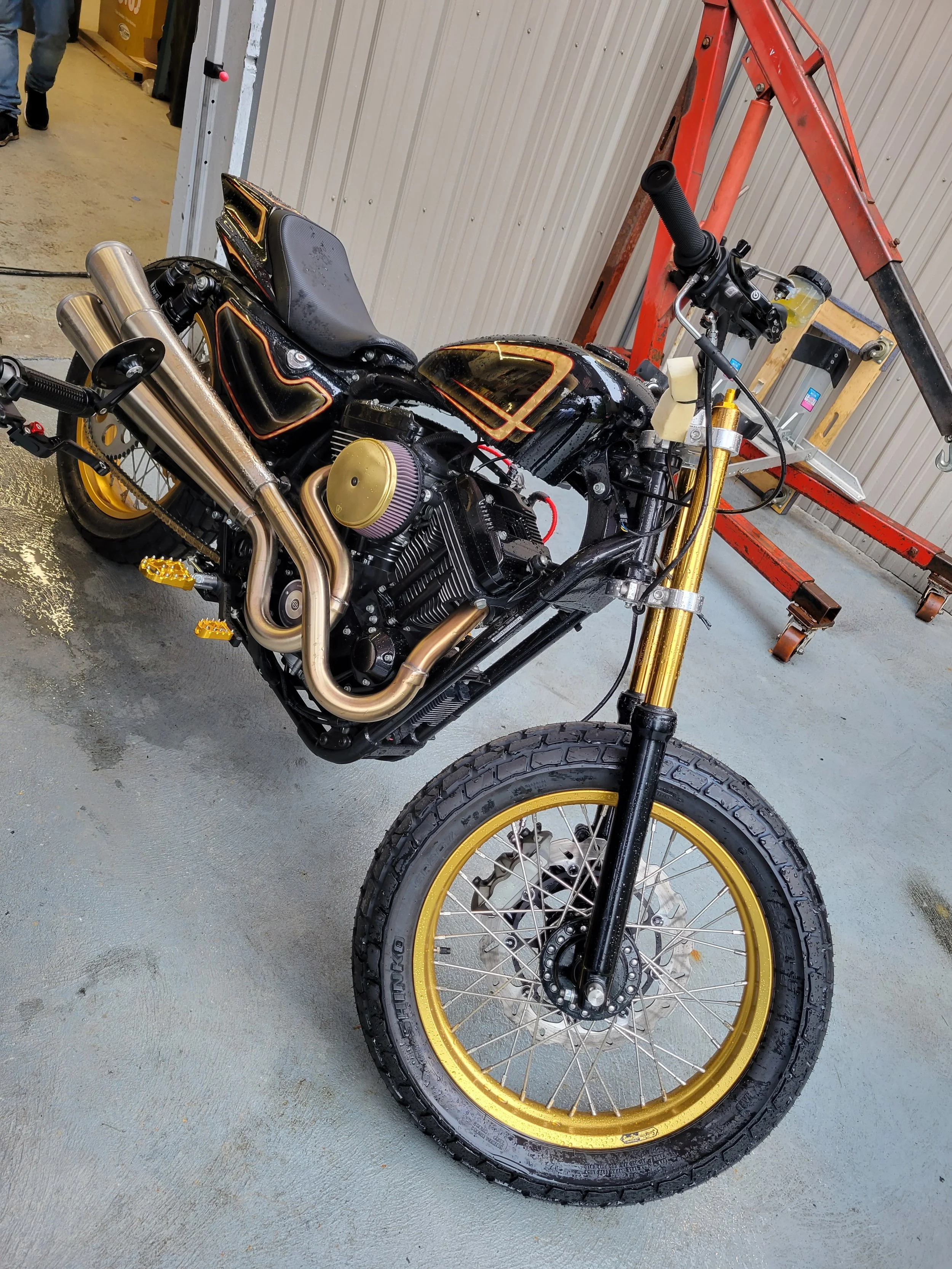 Black motorcycle with gold accents and gold wheels in a garage, with a red scaffold nearby.