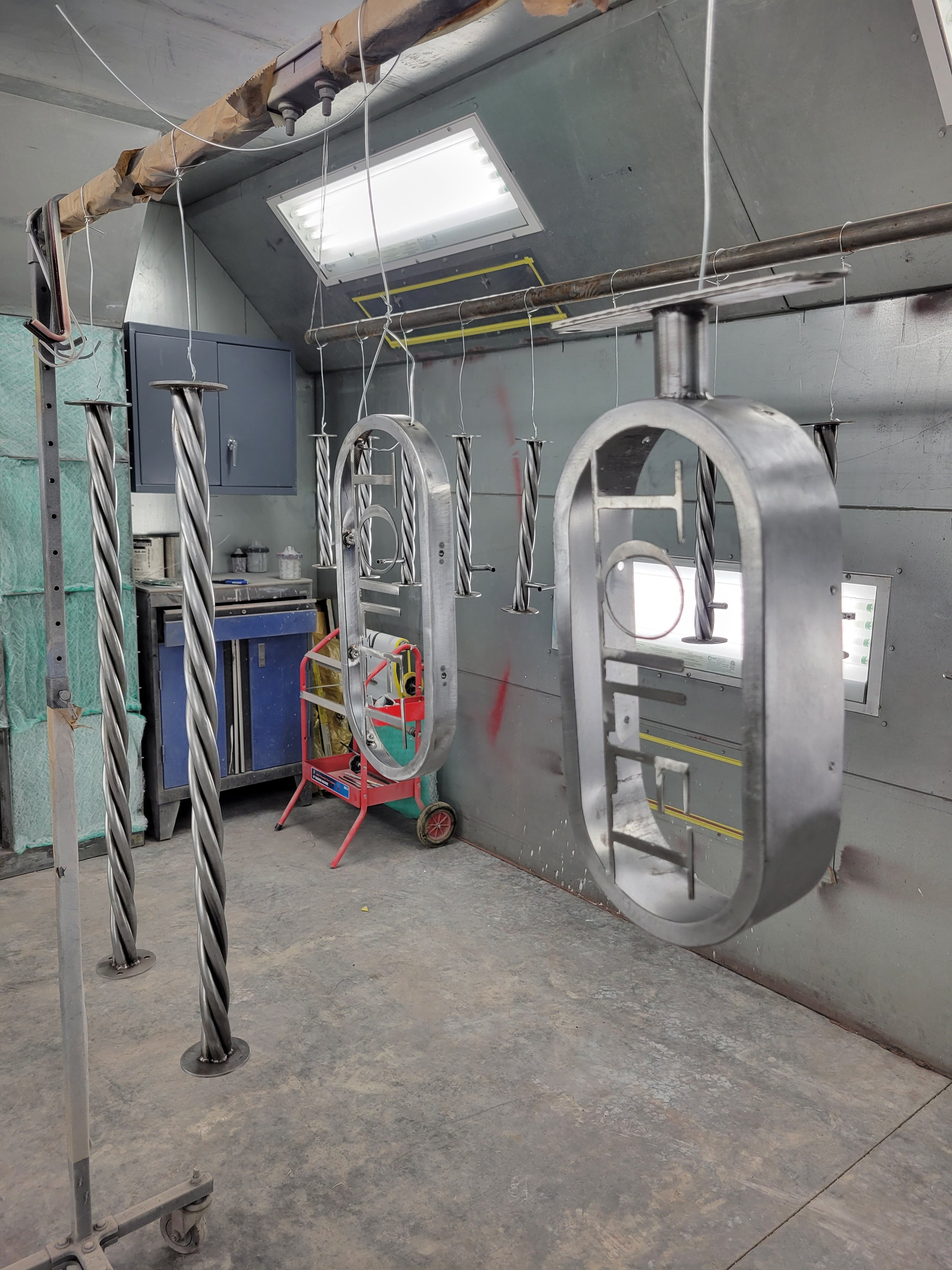 Metal structures hanging in a workshop, with drill bit-like tools attached. The background shows a grey wall, a blue cabinet, and various tools and supplies. The ceiling has fluorescent lighting panels and exposed piping.