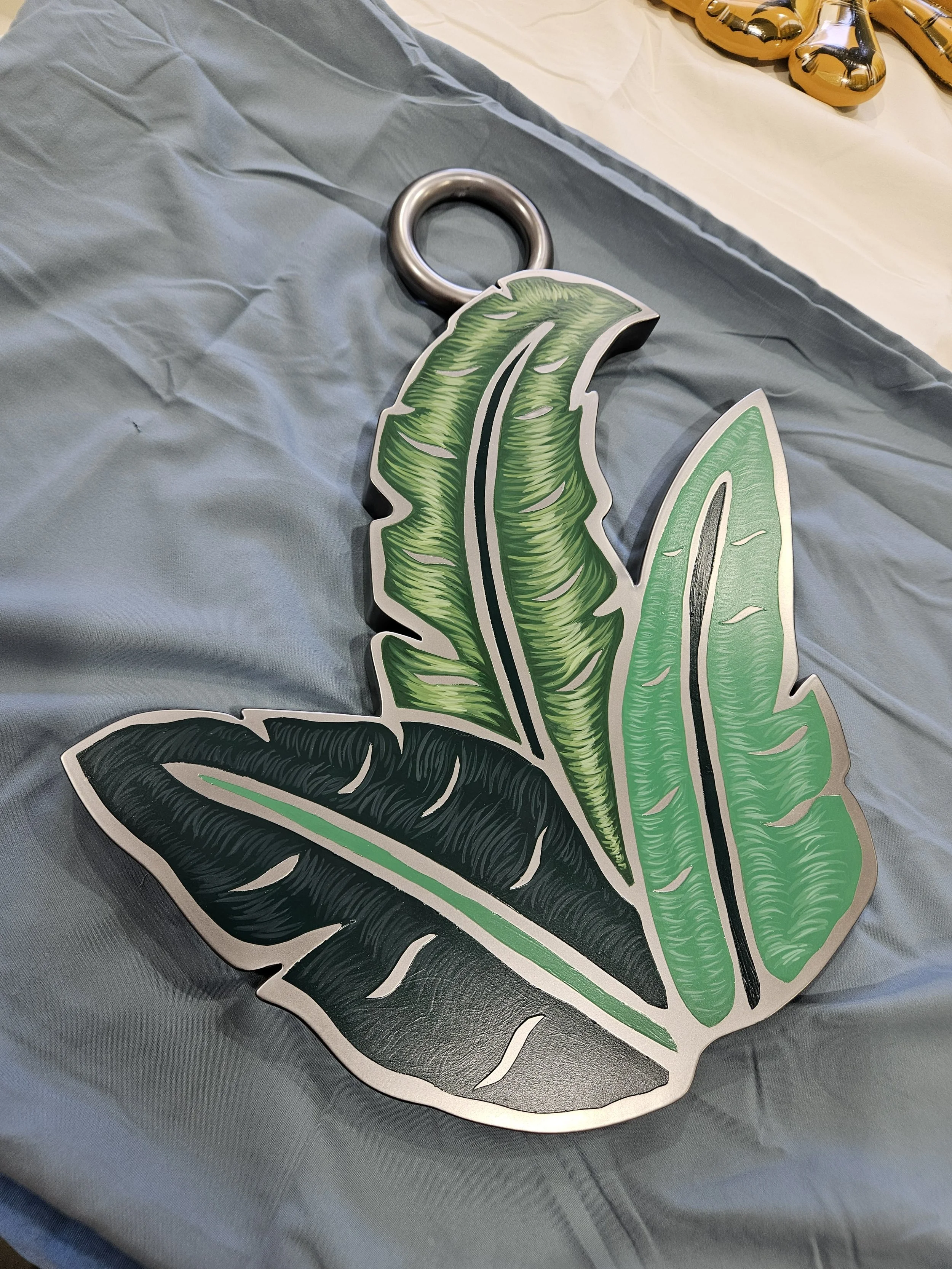Decorative sign in the shape of three green and black tropical leaves with a metal ring at the top, placed on a blue cloth table.