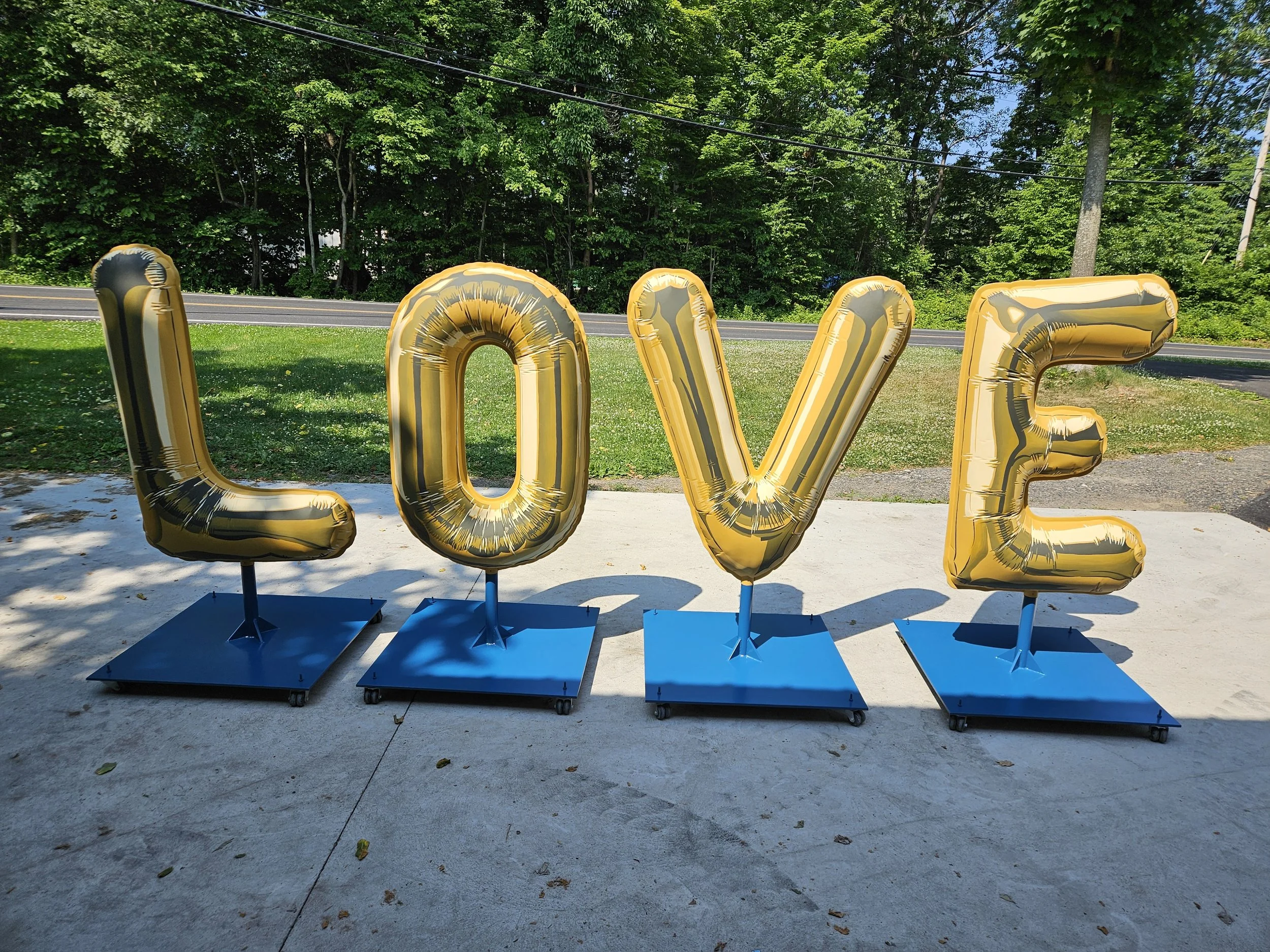 Large gold foil balloons spelling out the word "LOVE" on blue stands, outdoors on concrete with trees and a road in the background.
