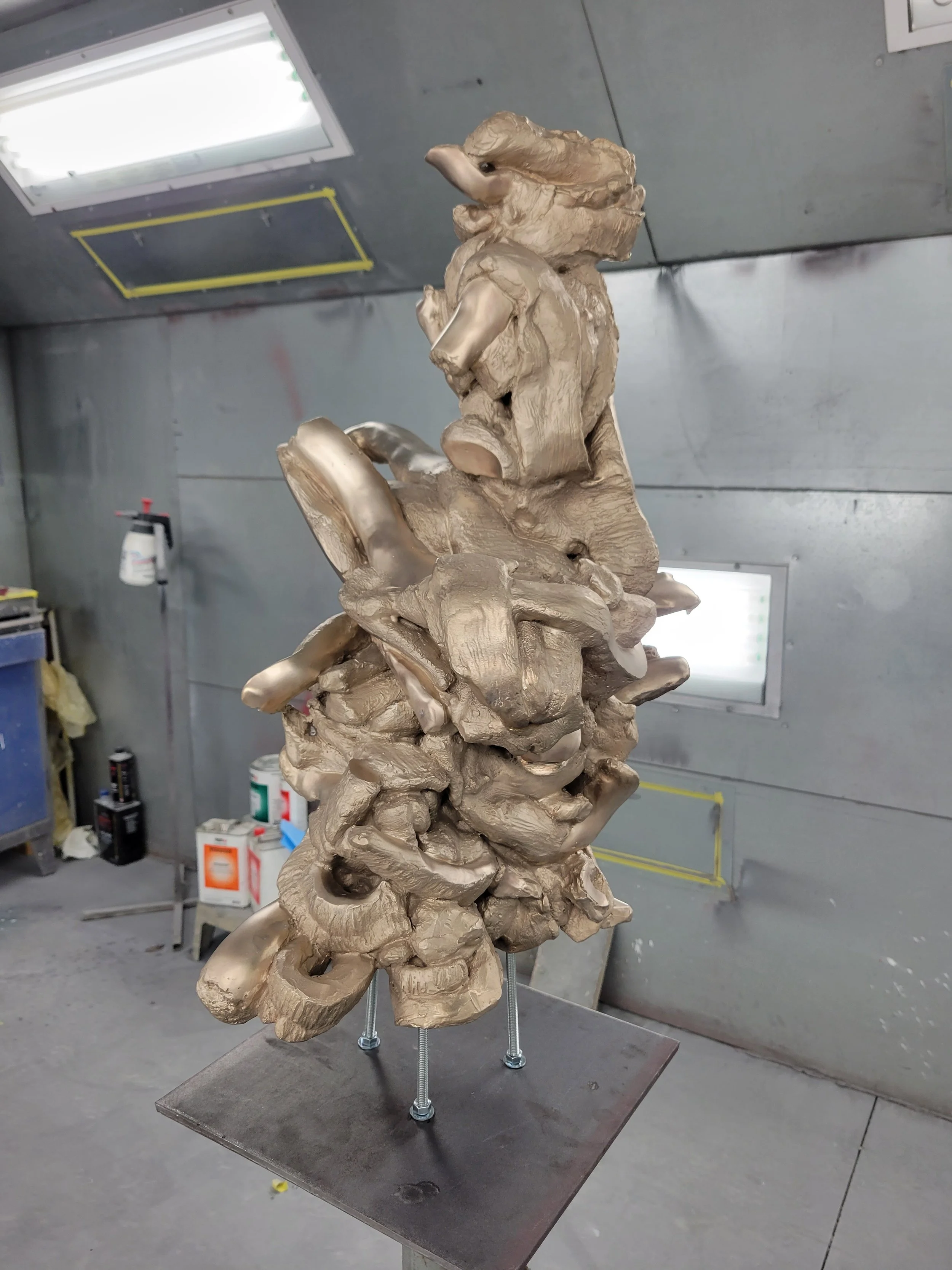 A modern abstract sculpture with organic, flowing shapes made of metallic or clay-like material, mounted on a metal base with four bolts, in an industrial workshop.
