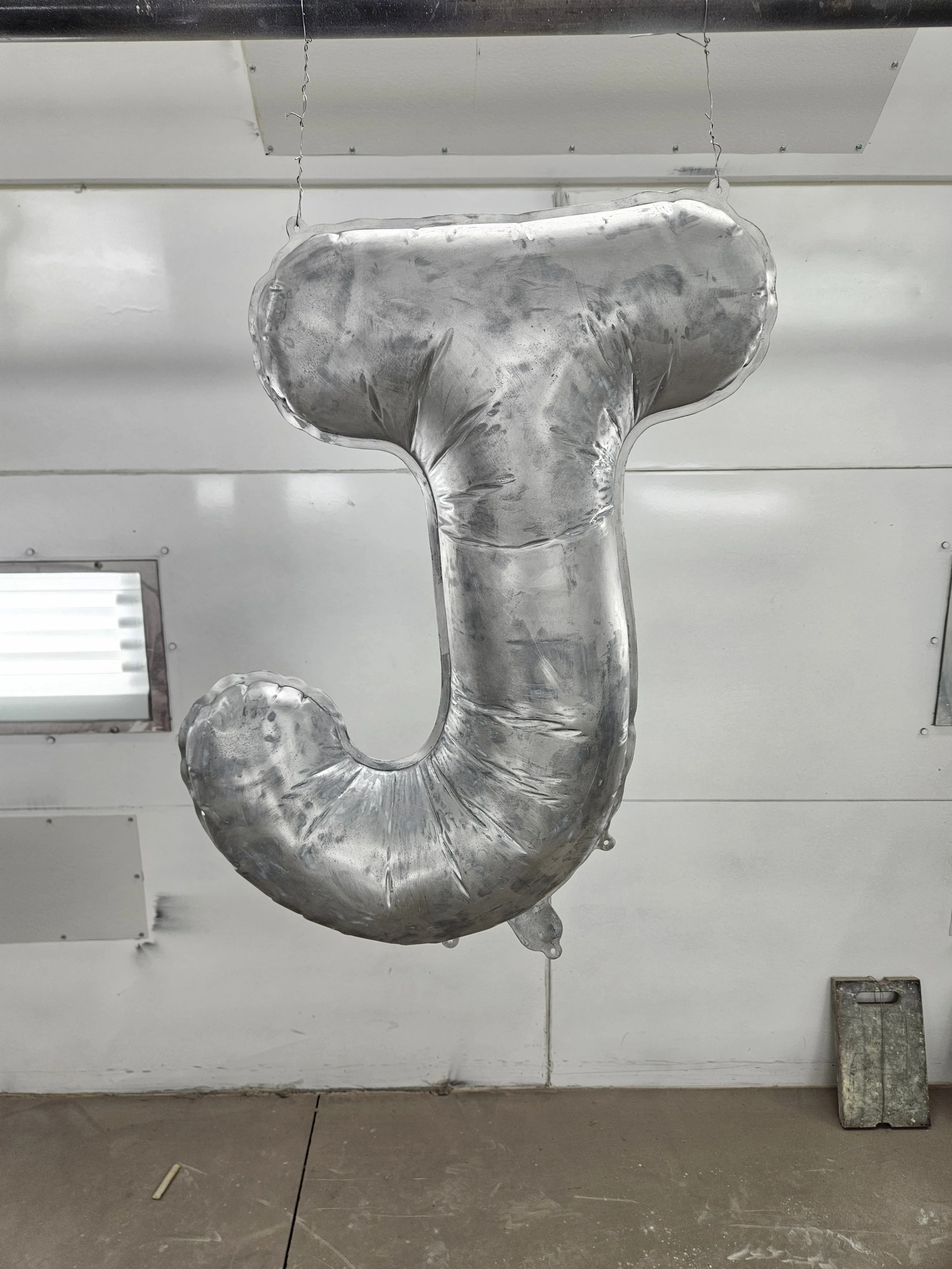 Silver foil balloon shaped like the letter J, hanging from the ceiling in an industrial or workshop space.