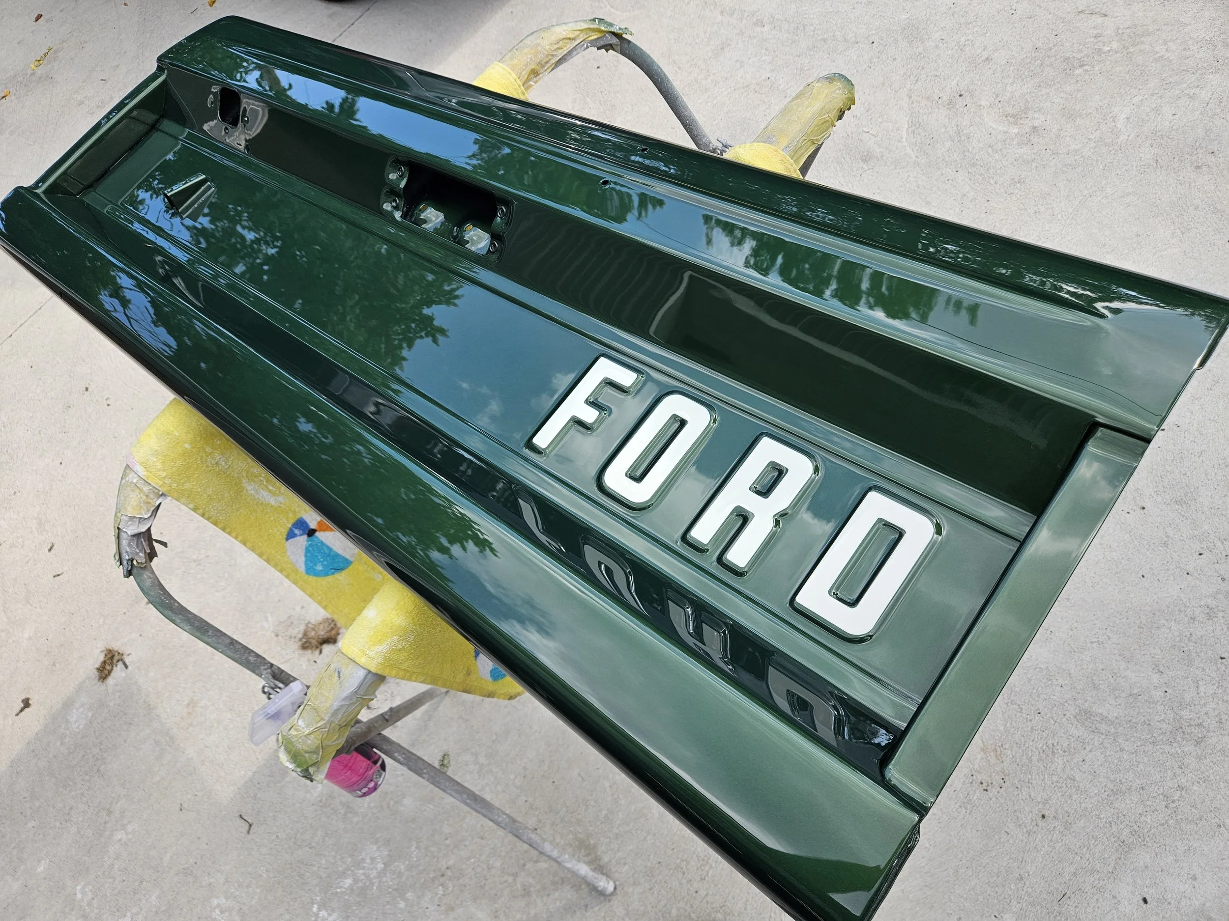 A painted green Jeep hood with the word "FORD" in white letters, resting on a yellow work stand outdoors.