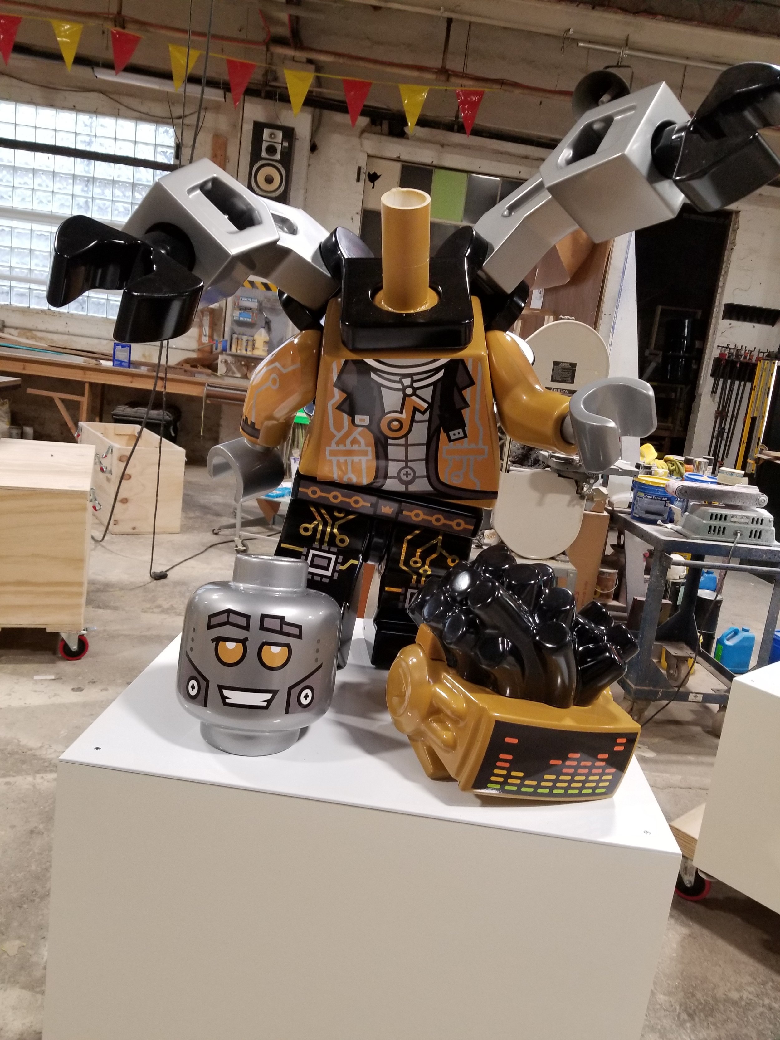A large robot sculpture made from LEGO-like materials, dressed in a black jacket and with an orange circuit pattern on the body. It has a cylindrical head, grey and black arms, and components resembling audio equipment on the base. The scene appears 