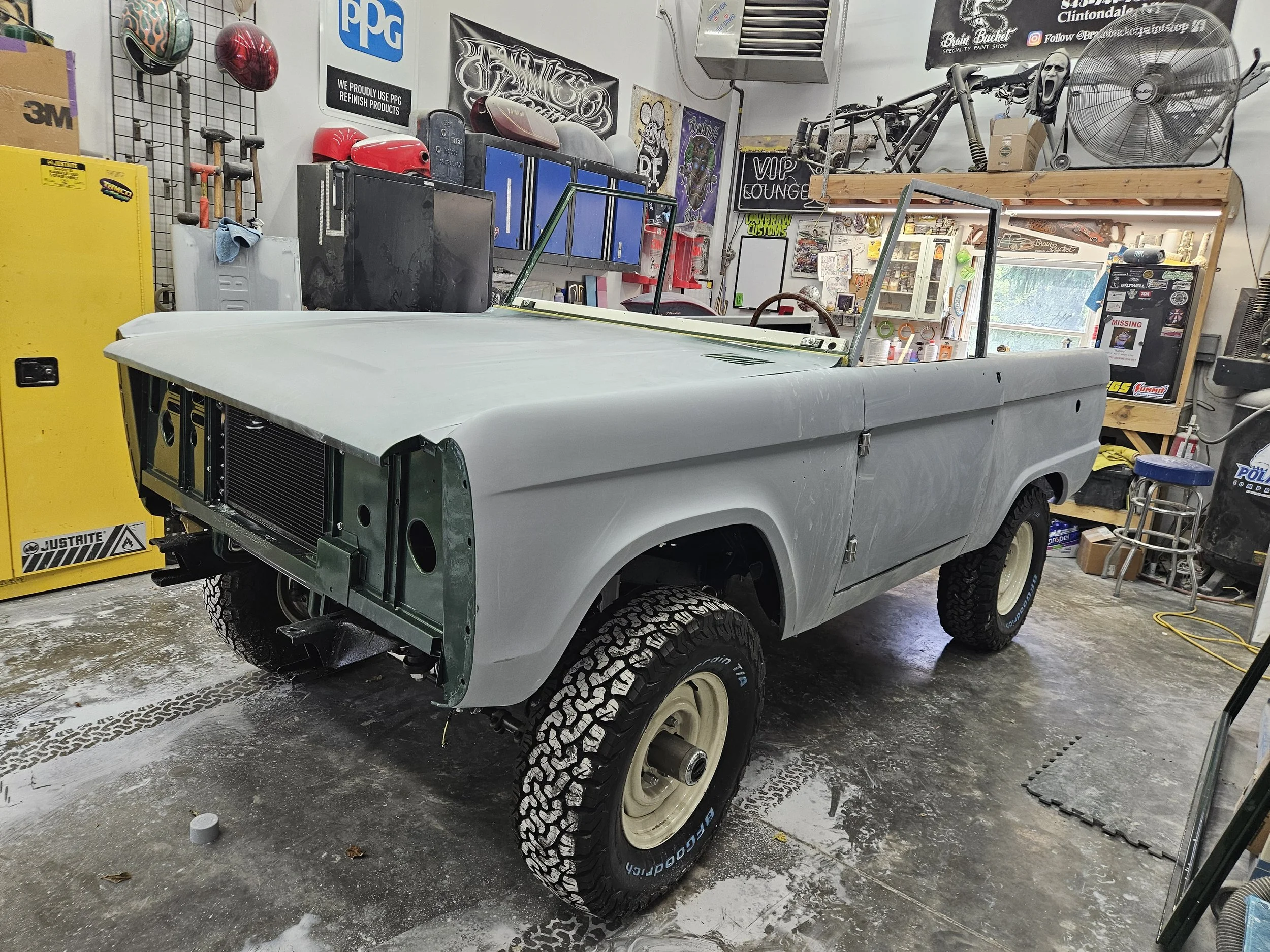 Unfinished vintage off-road vehicle in a garage, with a gray body, large off-road tires, and an open windshield frame, surrounded by various tools and equipment.