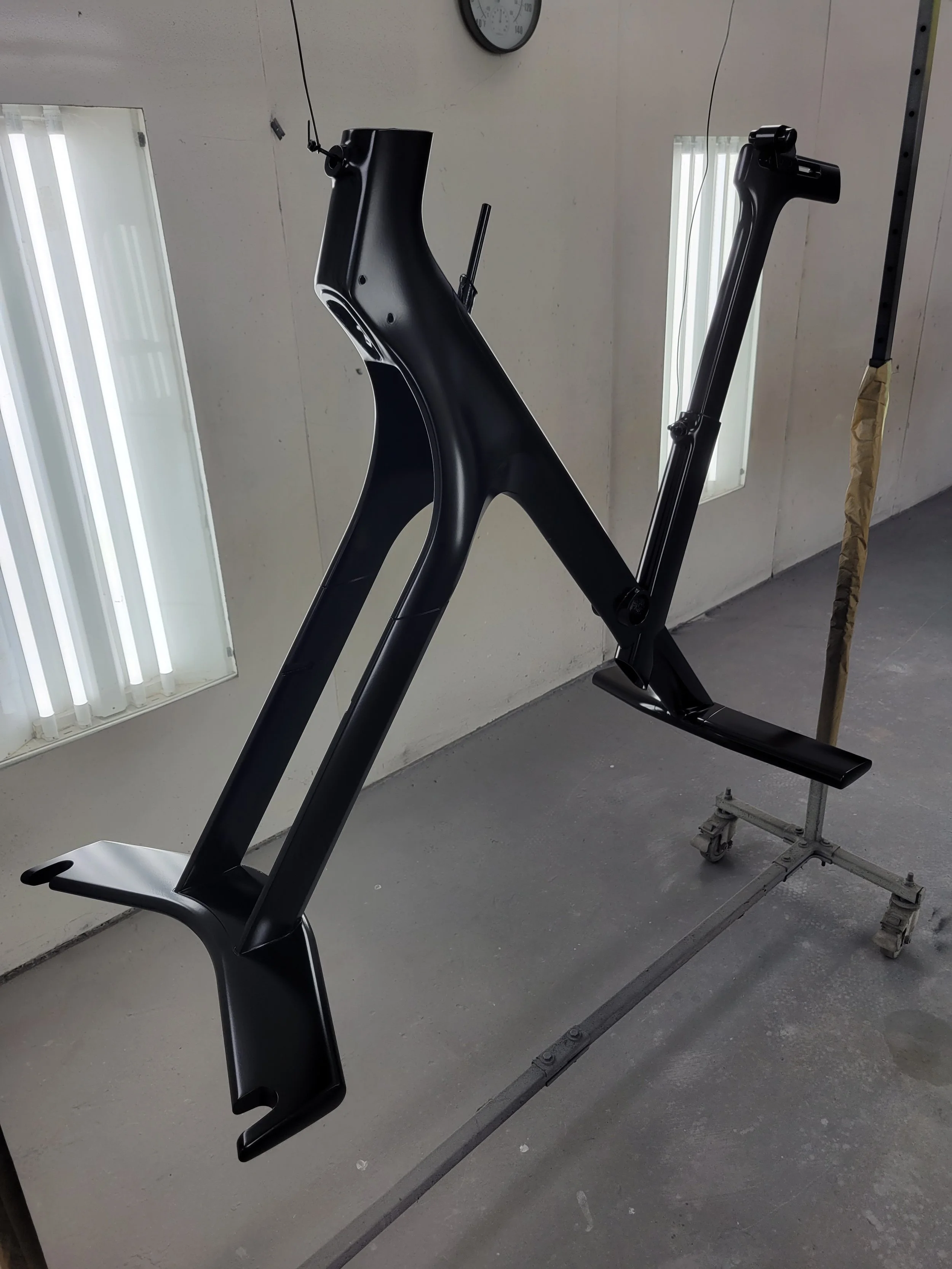 A black bicycle frame mounted on a work stand inside a room, with fluorescent lights and a clock on the wall.