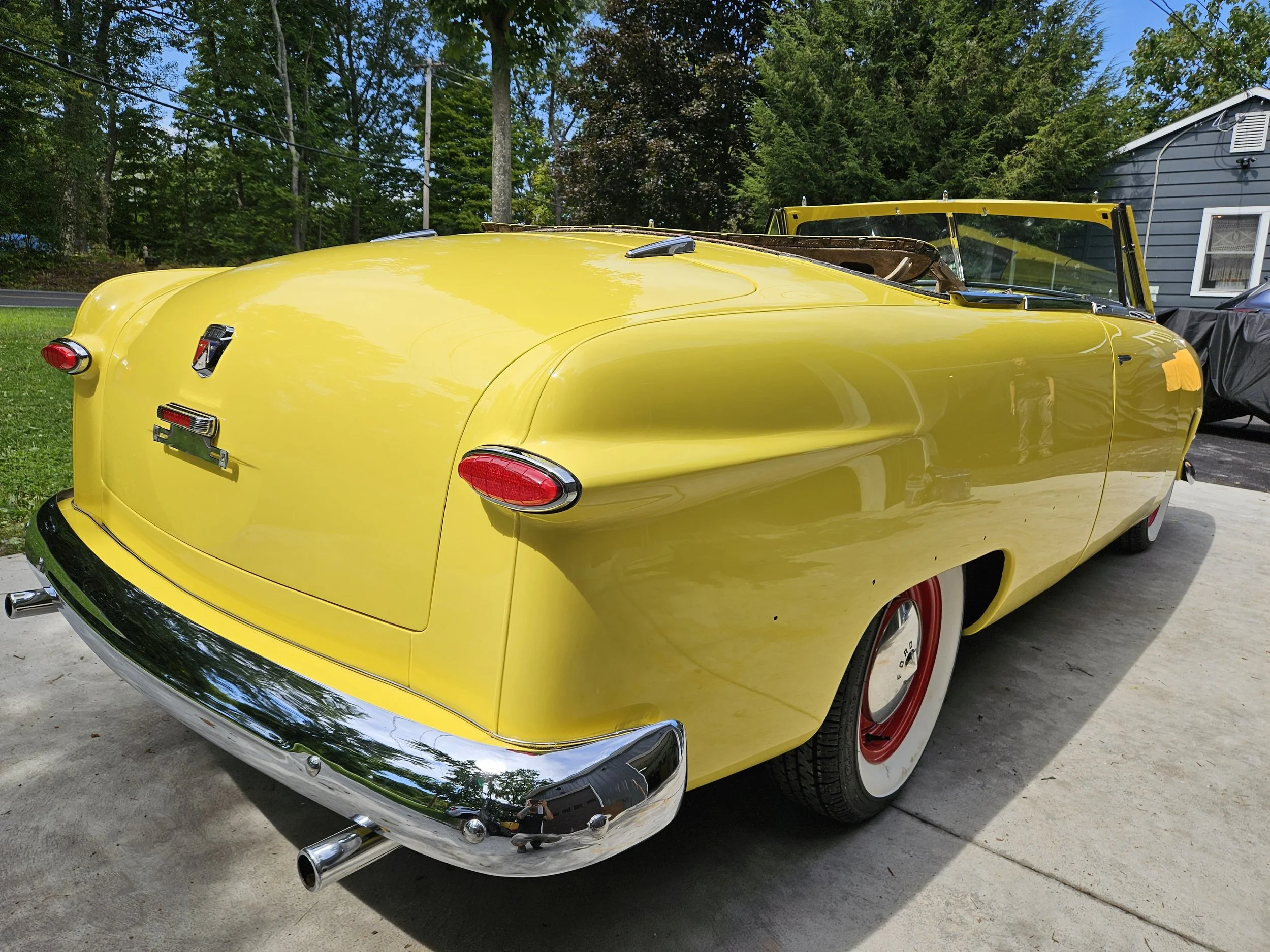 A vintage yellow convertible car with chrome bumper, whitewall tires, and red hubcaps parked on a concrete driveway.