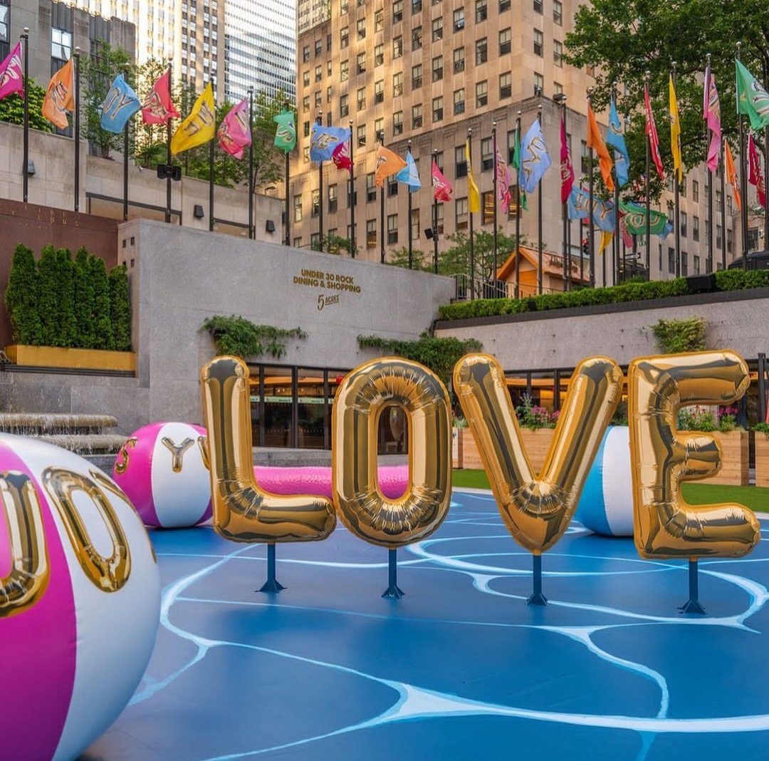 Golden balloon letters spelling LOVE on a blue court, surrounded by pink, white, and blue beach balls decorated with gold letters, outdoors in an urban setting with buildings and colorful flags above.