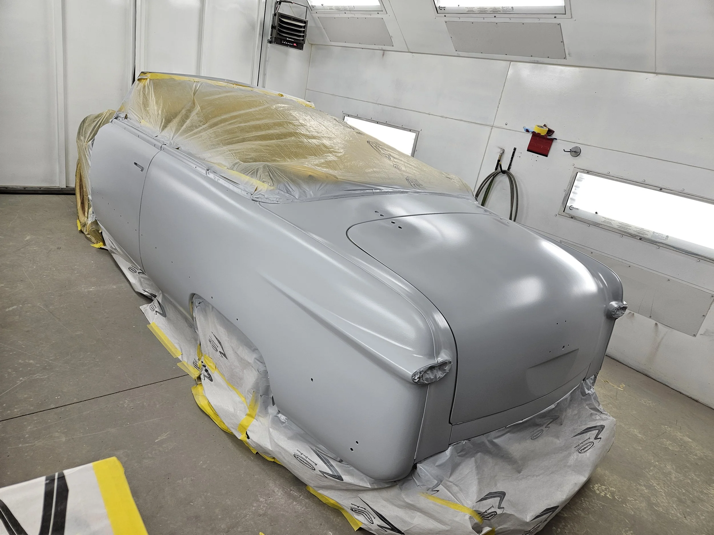 A classic car body in a paint booth, partially covered with masking paper and ready for painting, with a sleek silver finish.
