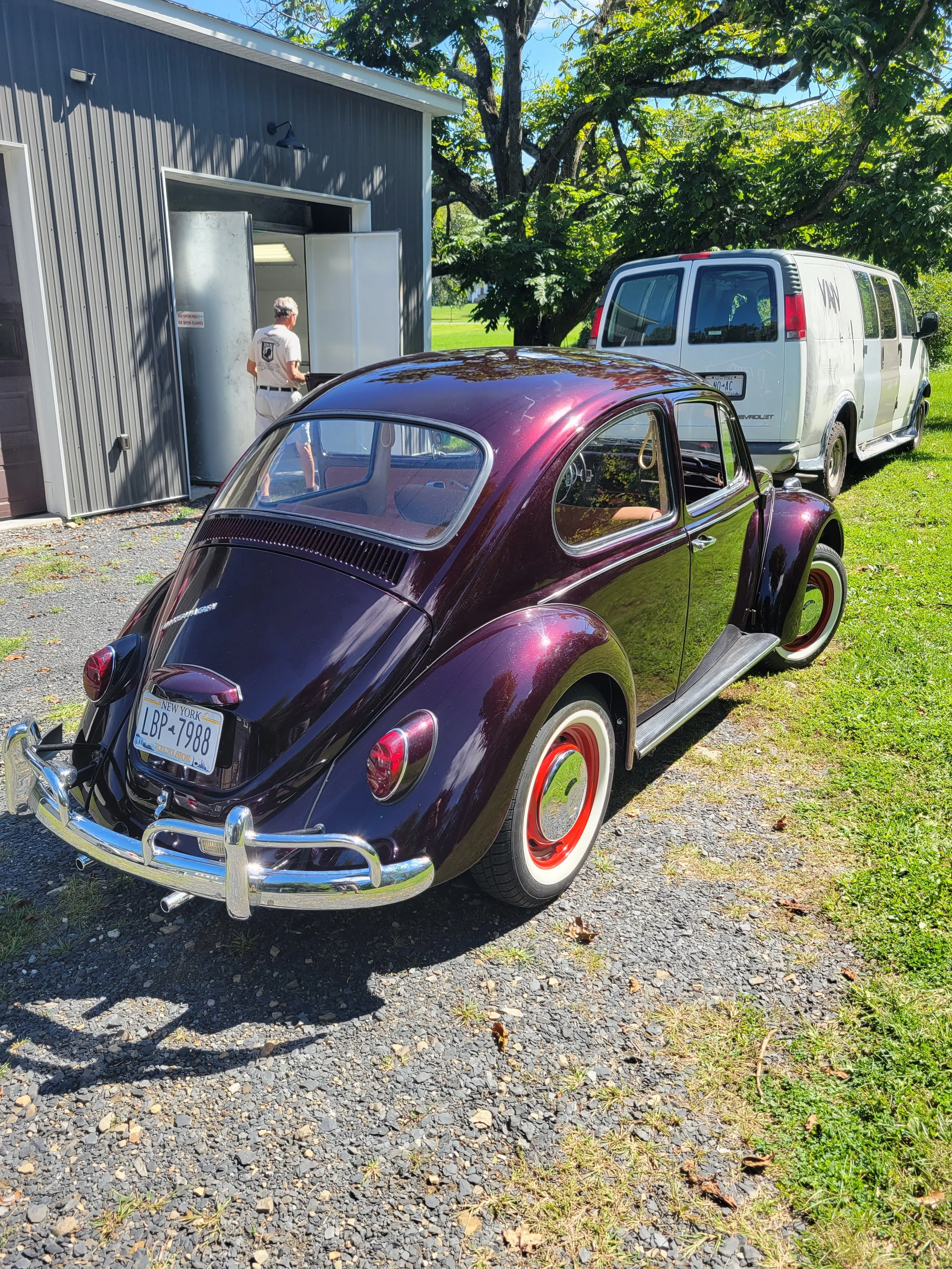 A vintage purple Volkswagen Beetle with white and red wheel rims parked on a gravel area near a grassy patch, against a backdrop of green trees and a building with an open garage door. A person is seen near the garage, and a white van is parked nearb