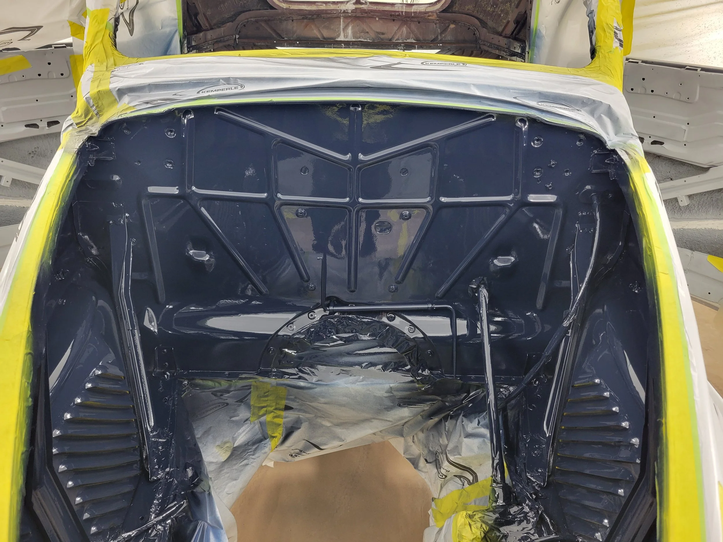 View of the inside of a car's engine bay, freshly painted in dark blue, with areas masked off with masking tape and paper for painting.