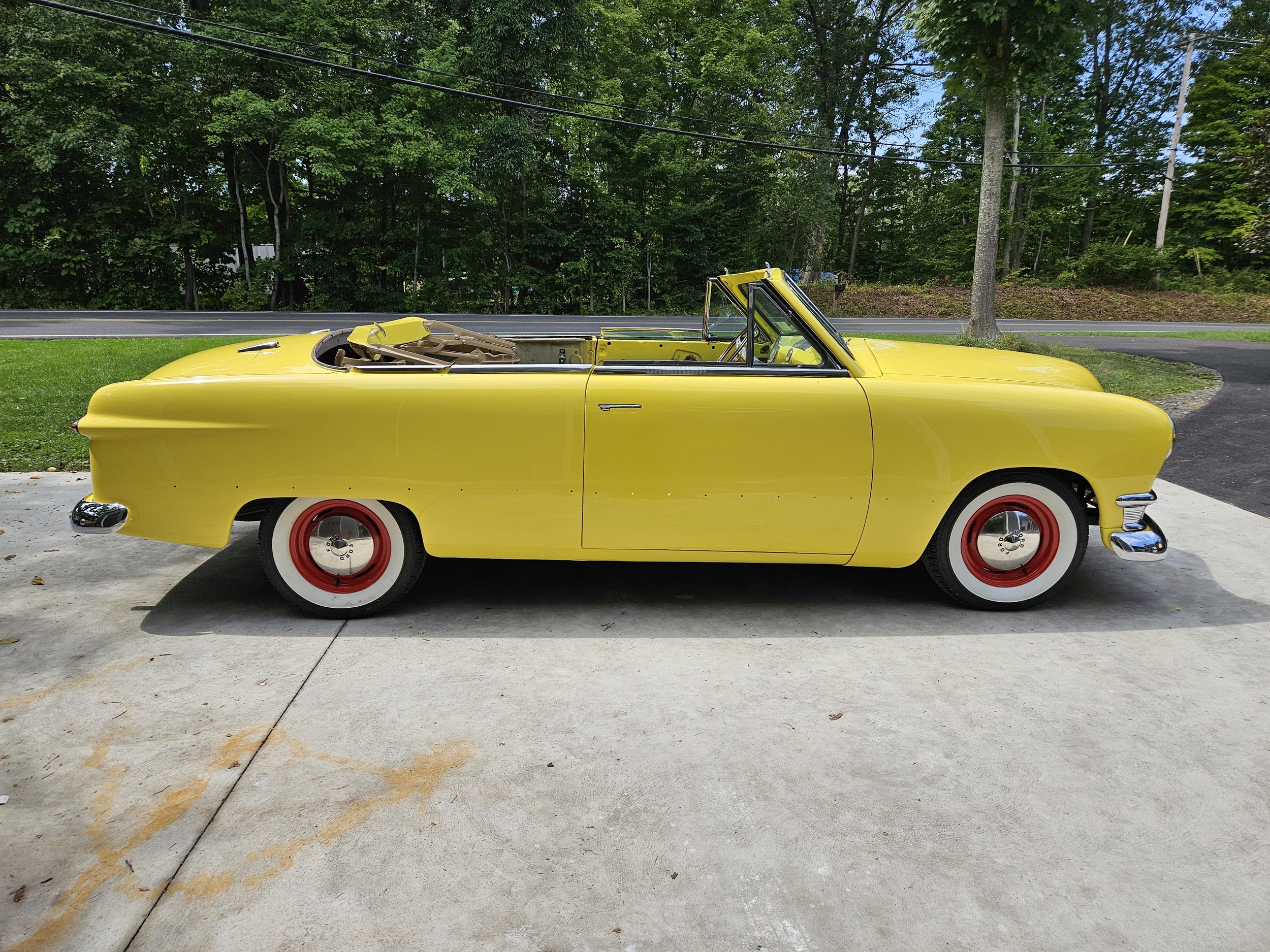A vintage yellow convertible car with whitewall tires parked on a concrete driveway with a grassy yard and trees in the background.