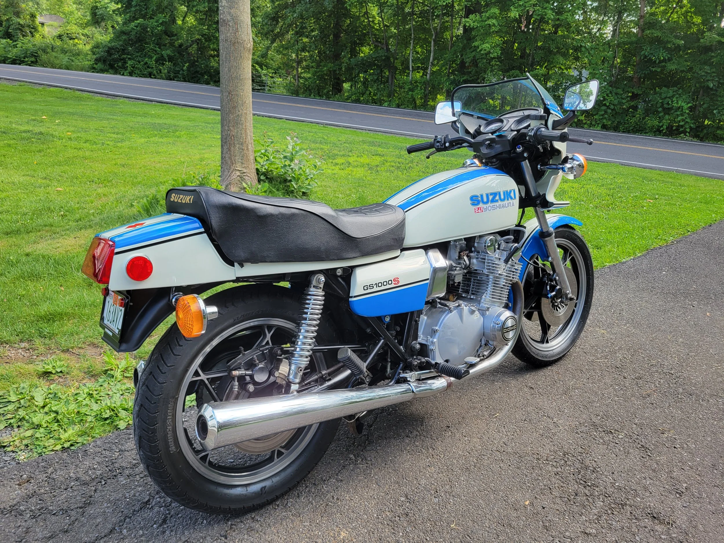 A classic Suzuki motorcycle parked on a paved path next to a grassy area with a tree and a road in the background.