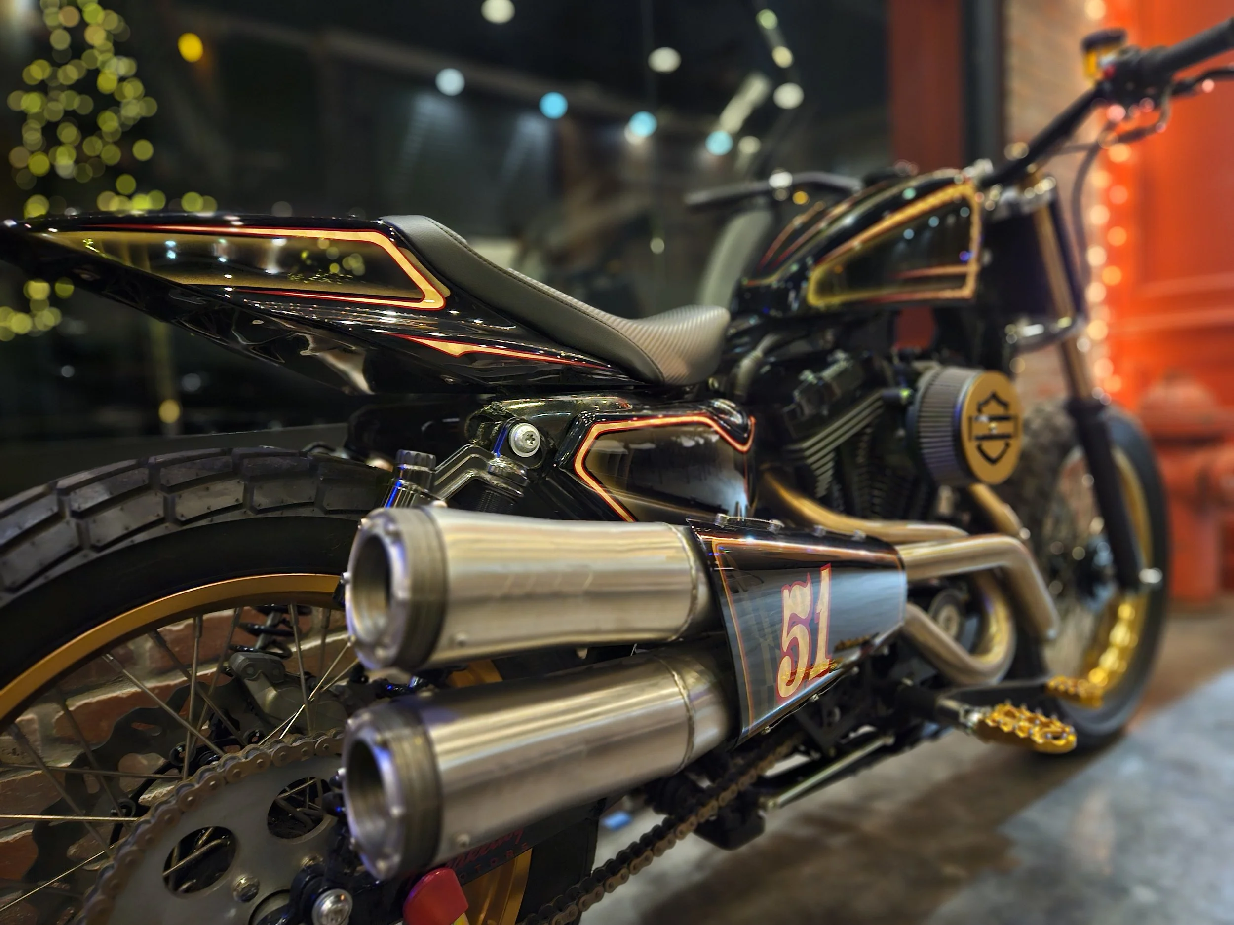 Close-up of a custom black and gold motorcycle with dual exhaust pipes, racing number 51, and yellow accents, displayed indoors.