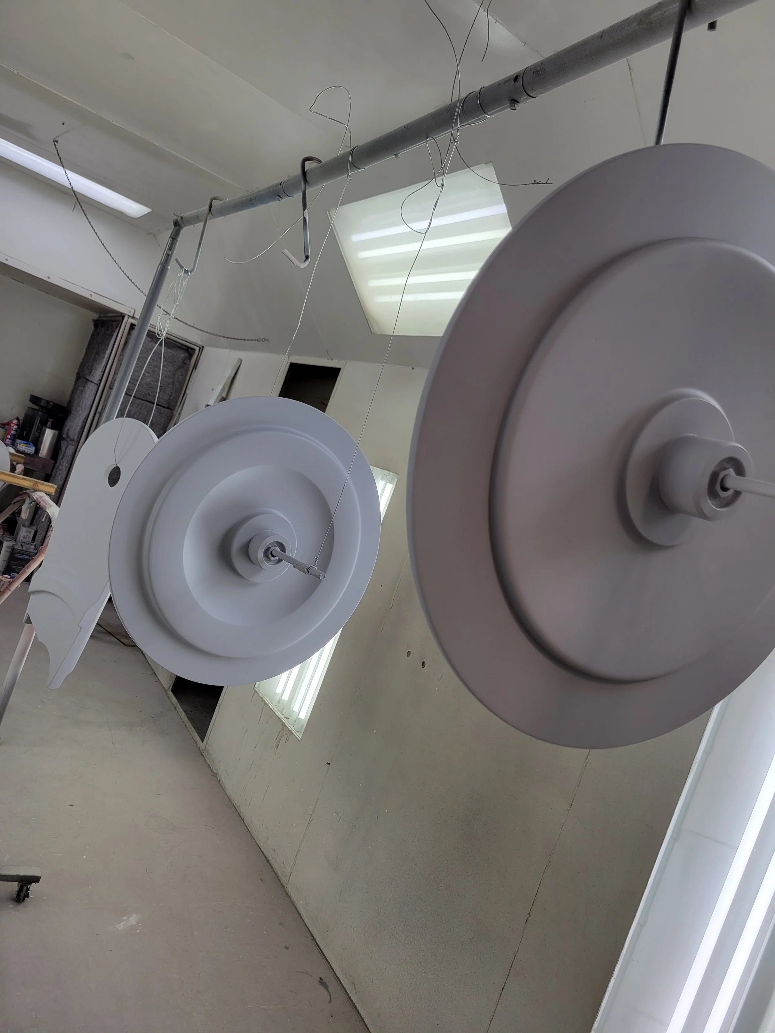 Two large white circular objects hanging from a metal pipe in a workshop or studio, with wires attached to them.