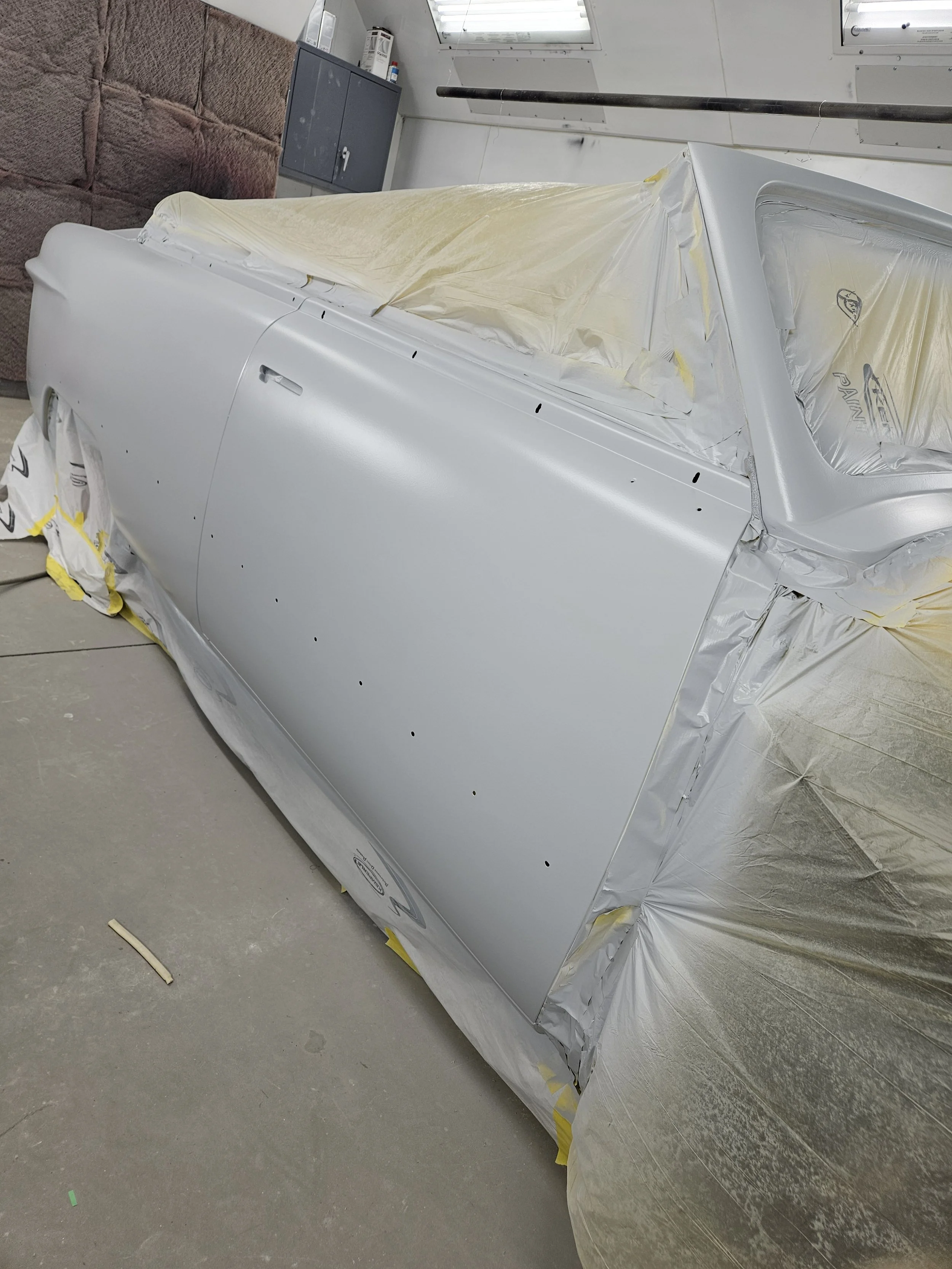 A silver car door being painted, covered with masking tape and paper for painting process in a garage or workshop.
