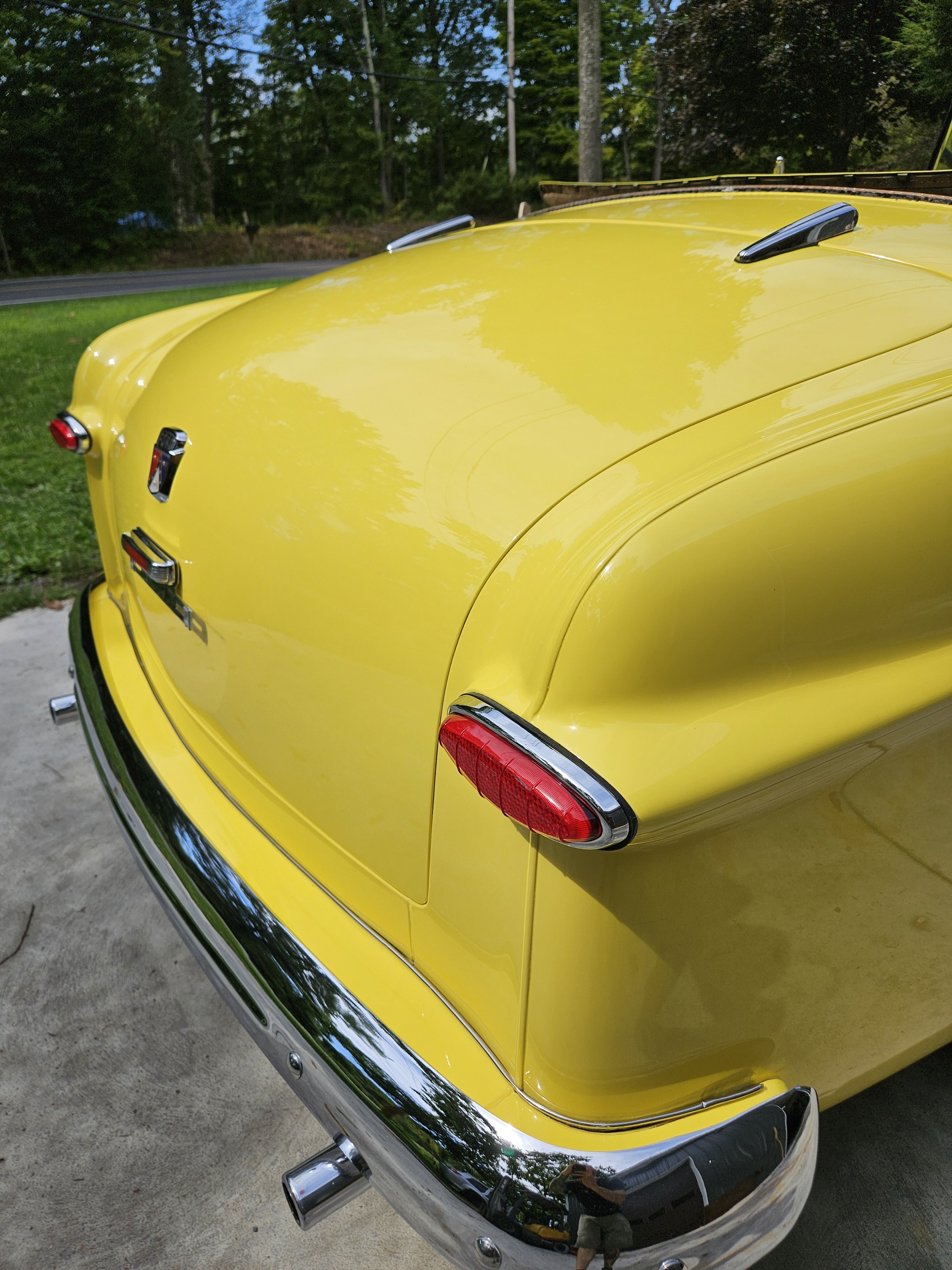Close-up of the rear end of a vintage yellow car with chrome bumper, tail lights, and side mirror, parked on concrete outside with green trees and grass in the background.