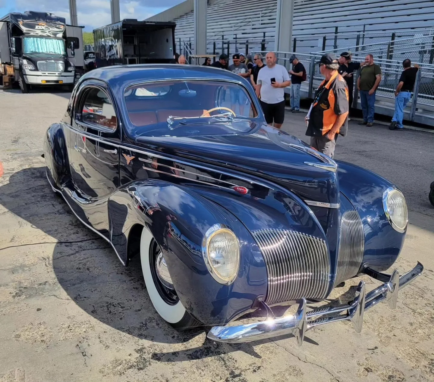A vintage black car with chrome accents and white-wall tires parked on a concrete surface, with people in the background at an outdoor event or car show.