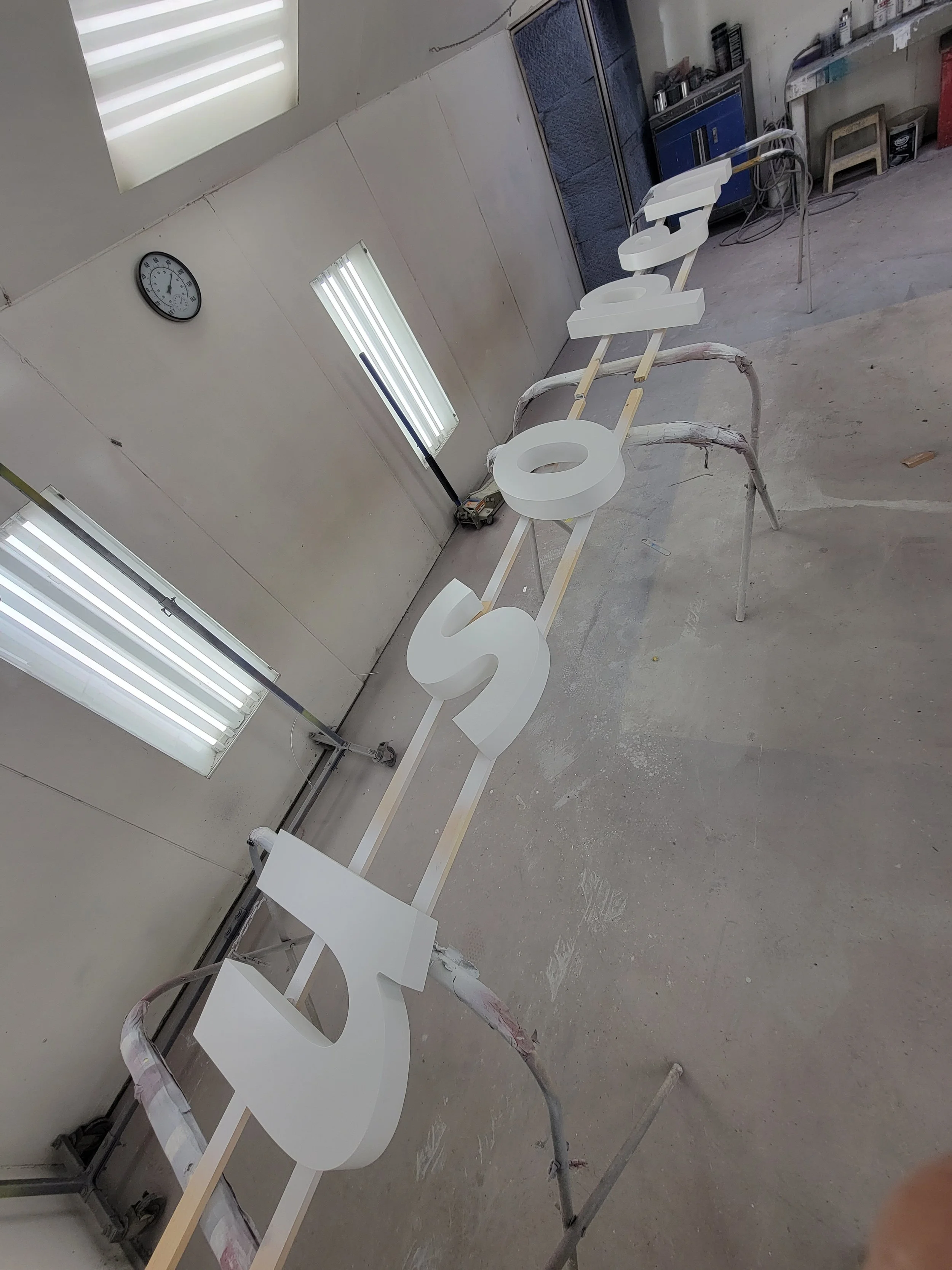 Large white letters spelling 'BESO' placed on wooden supports in an indoor workshop with fluorescent lights, clock, and storage cabinets.