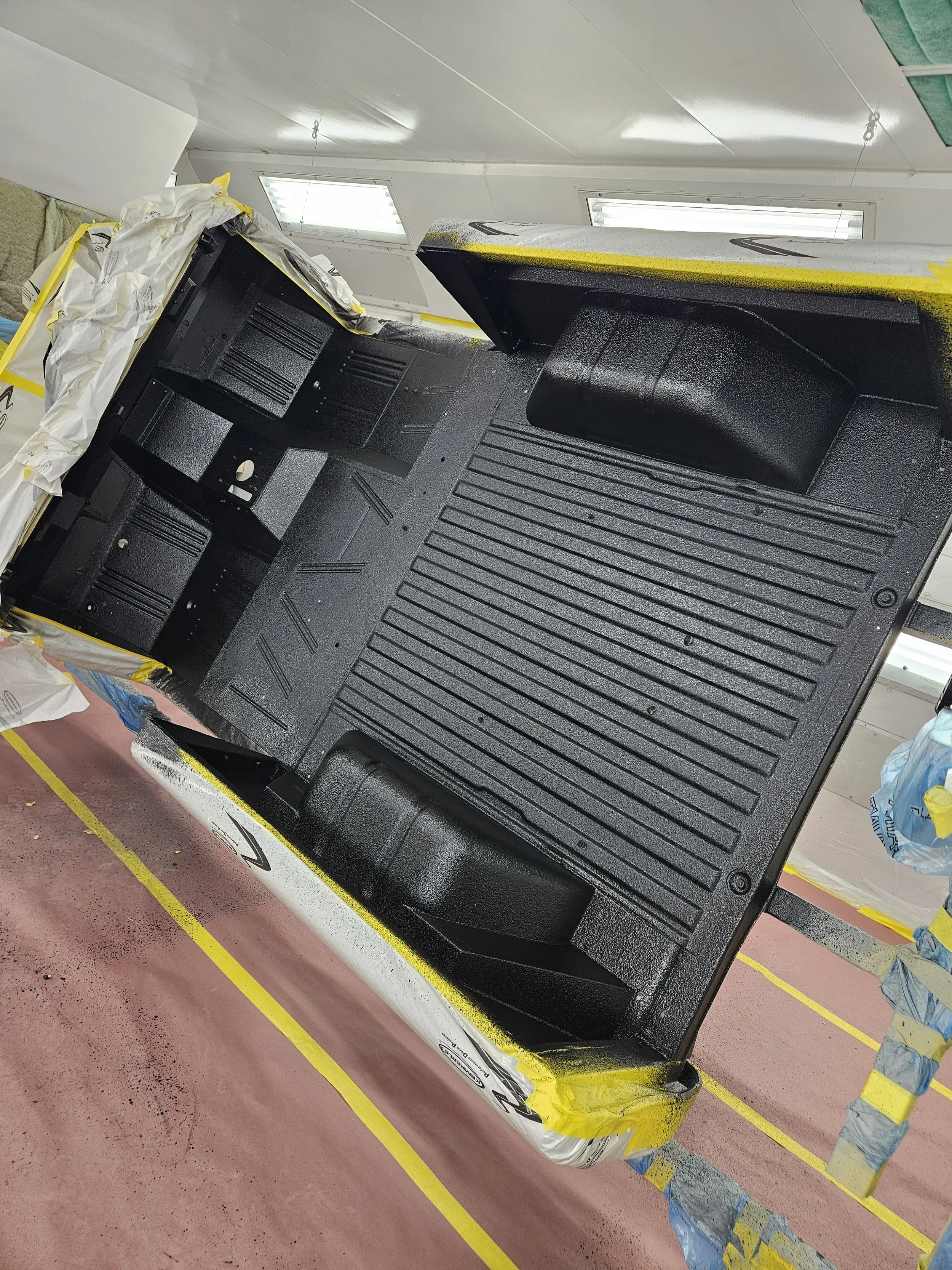 Interior of a vehicle's cargo area being painted black, with masking tape and plastic covers around the edges, in a well-lit workshop.