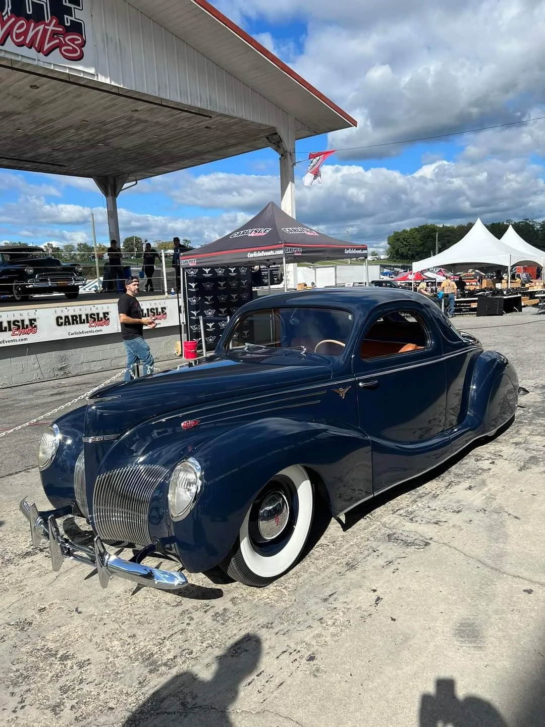 Vintage dark blue car with whitewall tires parked at a car show, with tents and people in the background under partly cloudy skies.