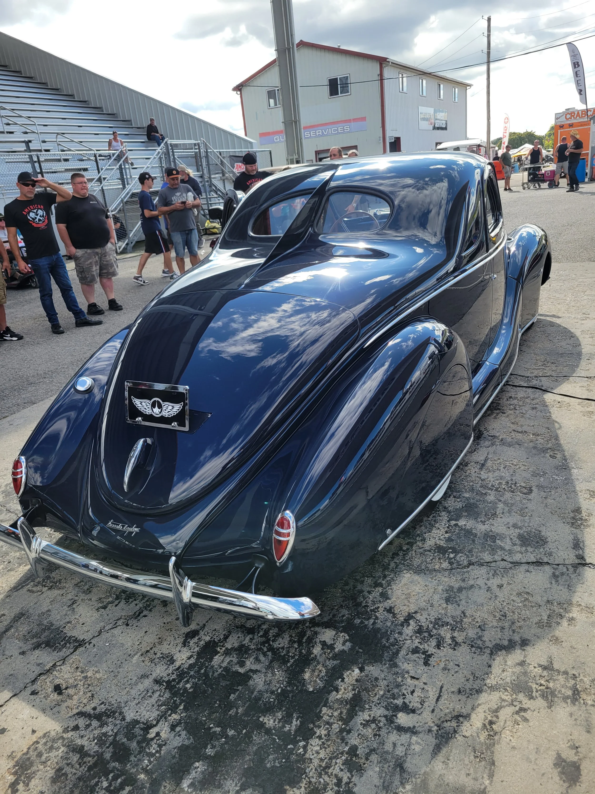 A vintage black luxury car with a streamlined design and chrome accents parked at a car show, with people gathered around and a building in the background.