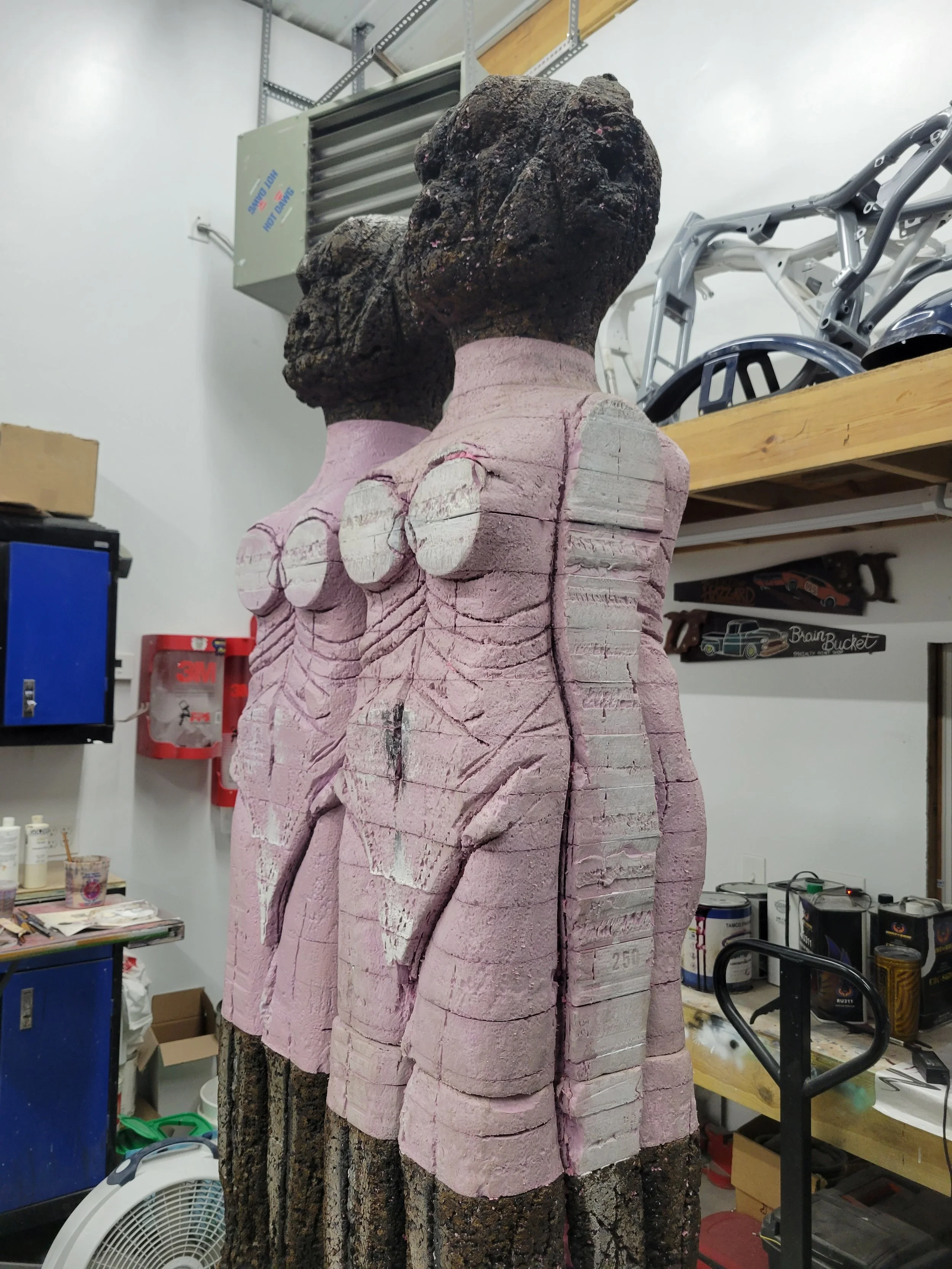 Carved wooden sculpture of three humanoid figures with dark heads and pink torsos, standing side by side in a workshop.