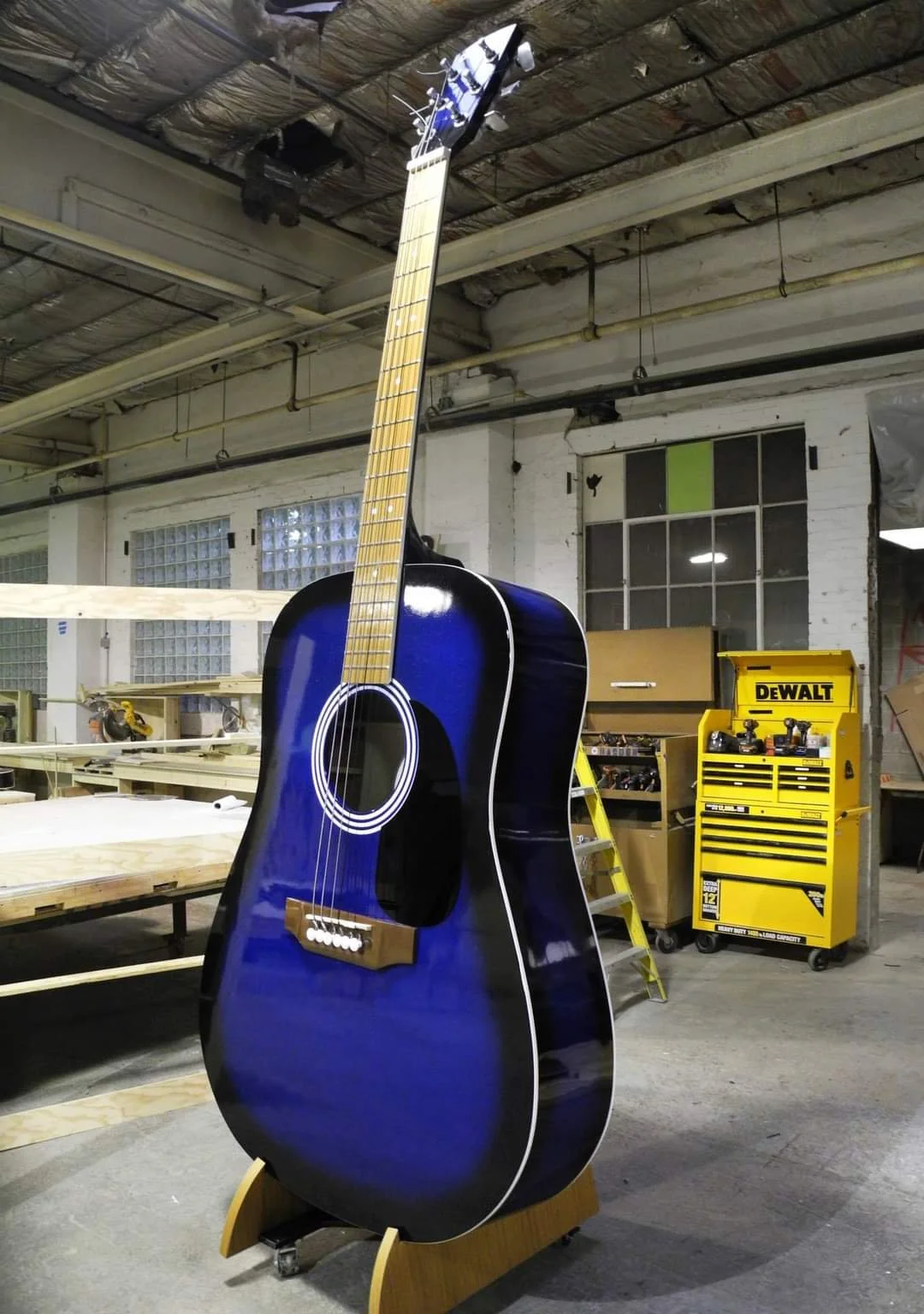 A blue acoustic guitar standing upright in a woodworking workshop, with woodwork tools and a yellow Dewalt tool chest in the background.